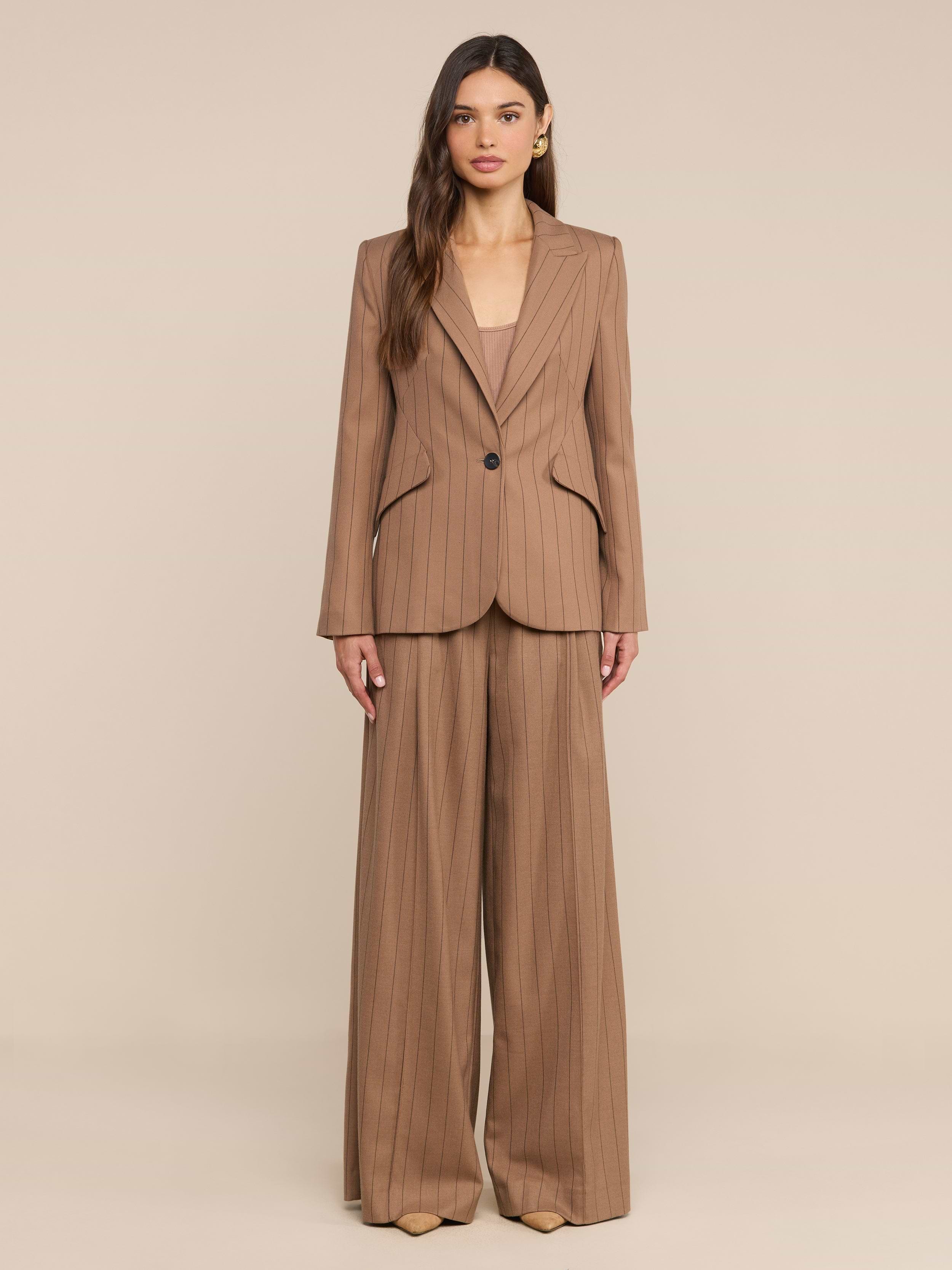 Une femme aux longs cheveux ondulés et au maquillage discret se tient debout, face à lobjectif, vêtue dun tailleur à fines rayures beige clair composé dun pantalon « Connie Drawstring » et dun blazer à un bouton, posant devant un fond beige uni.