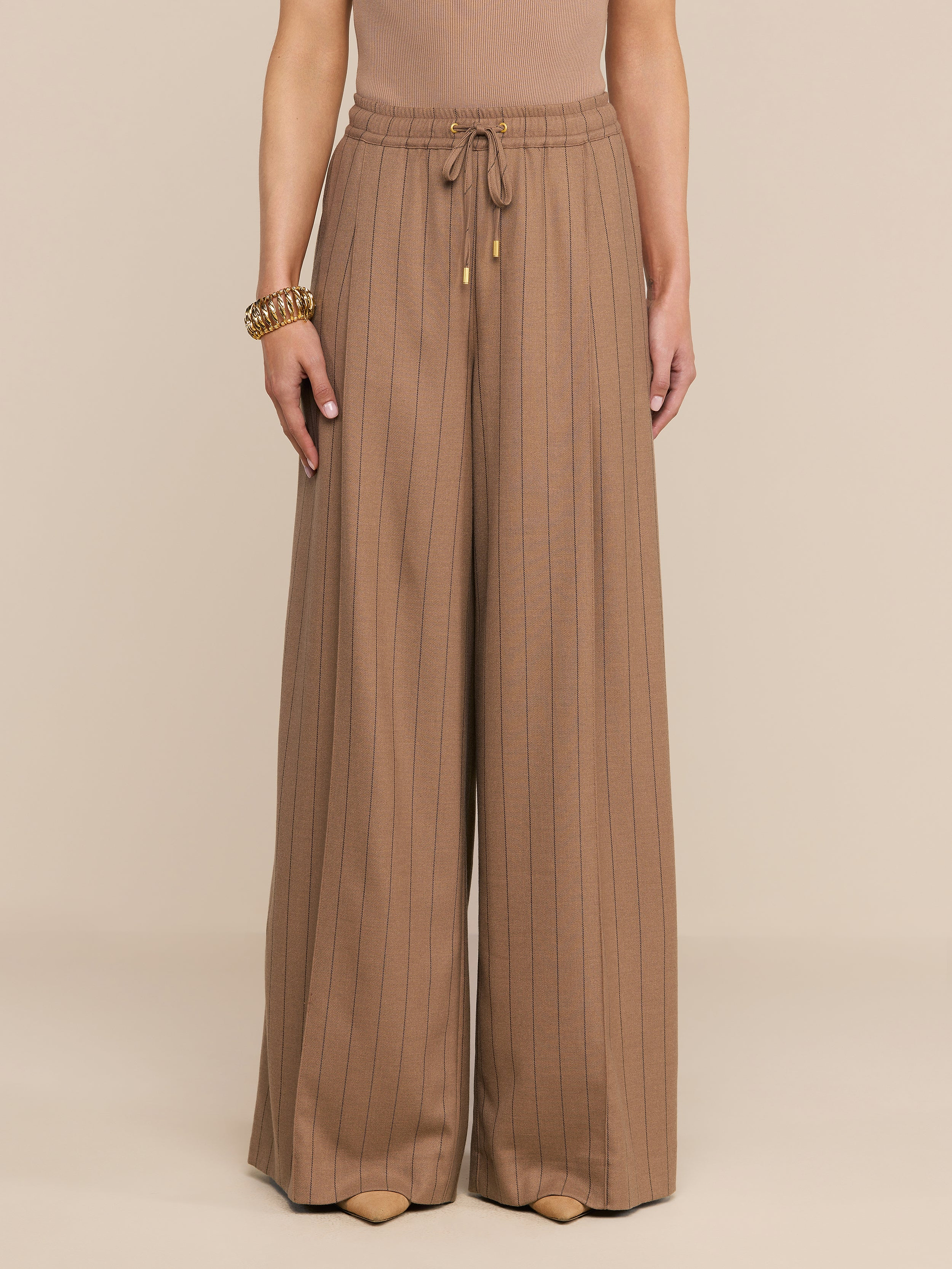 Mannequin portant le pantalon « Connie Drawstring » à fines rayures beiges, caractérisé par une taille haute, une coupe à jambes larges et un cordon de serrage à la taille, associé à un bracelet doré.