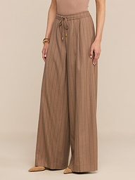 Une personne porte le pantalon « Connie Drawstring » — un pantalon à taille haute, à jambes larges et à fines rayures, doté d’un cordon de serrage à la taille — qu’elle associe à des chaussures beiges à bout pointu, un haut sans manches et un bracelet en or, le tout sur un fond neutre.