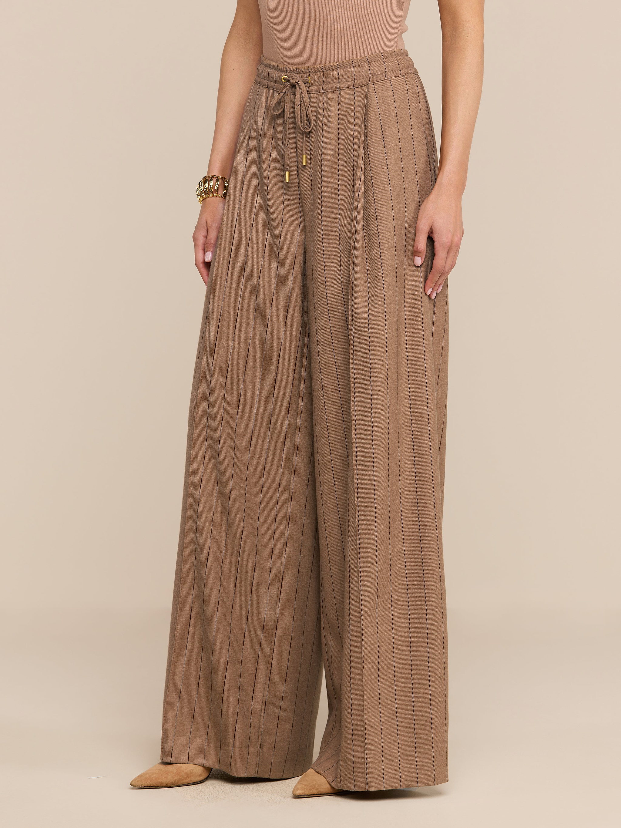 Une personne porte le pantalon « Connie Drawstring » — un pantalon à taille haute, à jambes larges et à fines rayures, doté d’un cordon de serrage à la taille — qu’elle associe à des chaussures beiges à bout pointu, un haut sans manches et un bracelet en or, le tout sur un fond neutre.