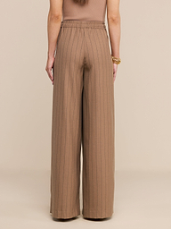Une femme, de dos, porte un pantalon « Connie Drawstring » — un pantalon à taille haute et à jambes larges, à fines rayures — ainsi quun haut sans manches. Ses bras sont détendus le long du corps ; elle porte un gros bracelet en or au poignet droit. Larrière-plan est beige.