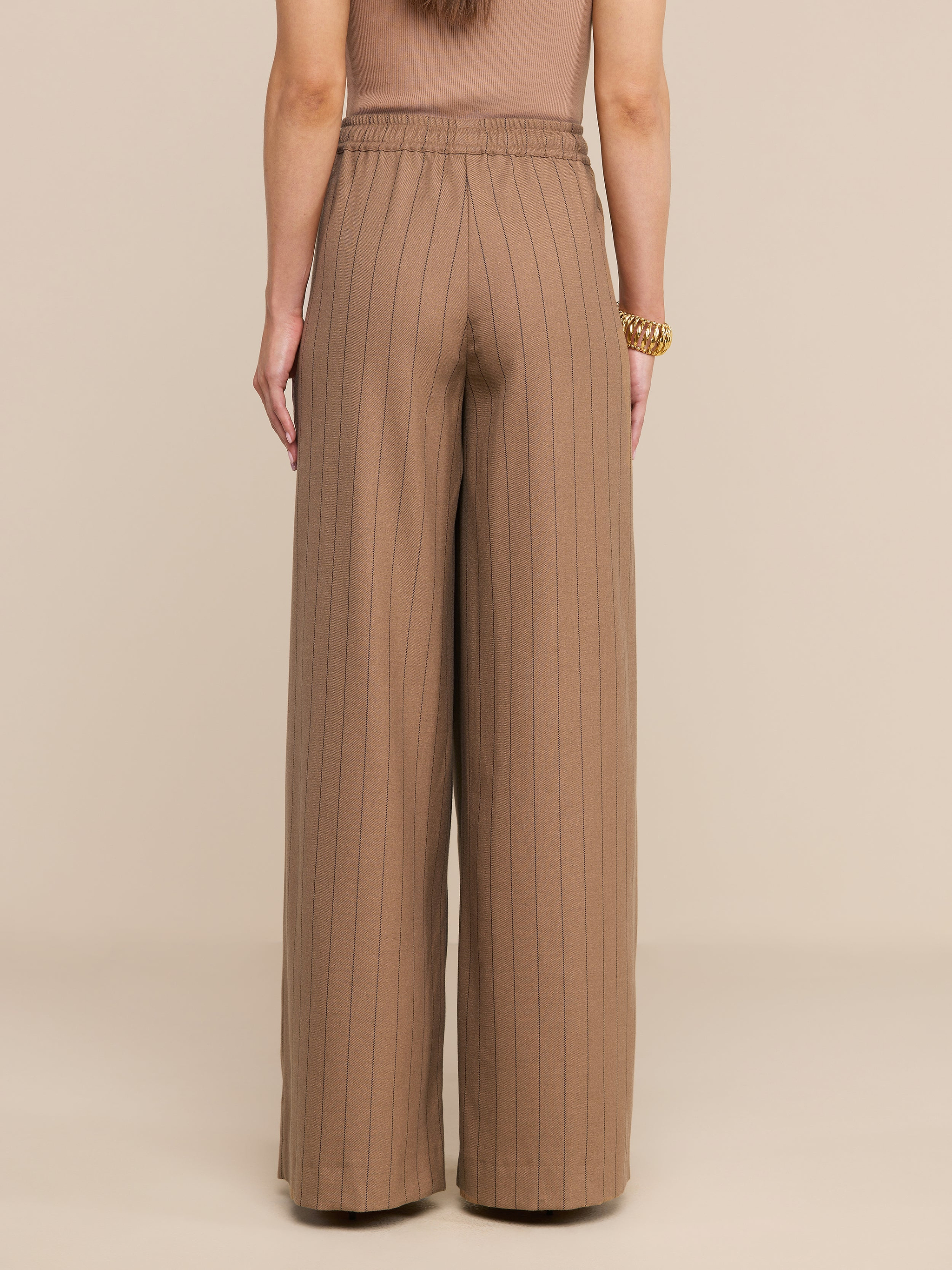 Une femme, de dos, porte un pantalon « Connie Drawstring » — un pantalon à taille haute et à jambes larges, à fines rayures — ainsi quun haut sans manches. Ses bras sont détendus le long du corps ; elle porte un gros bracelet en or au poignet droit. Larrière-plan est beige.