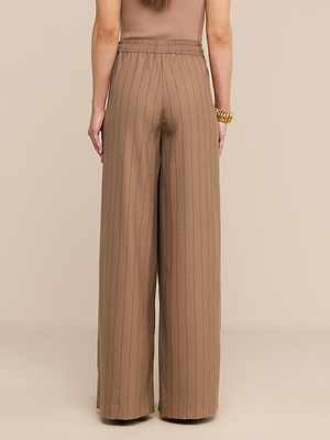 A woman stands facing away in Connie Drawstring Trousers—high-rise, wide-leg pinstripe pants—with a sleeveless top. Her arms are relaxed at her sides, wearing a chunky gold bracelet on her right wrist. The background is beige.
