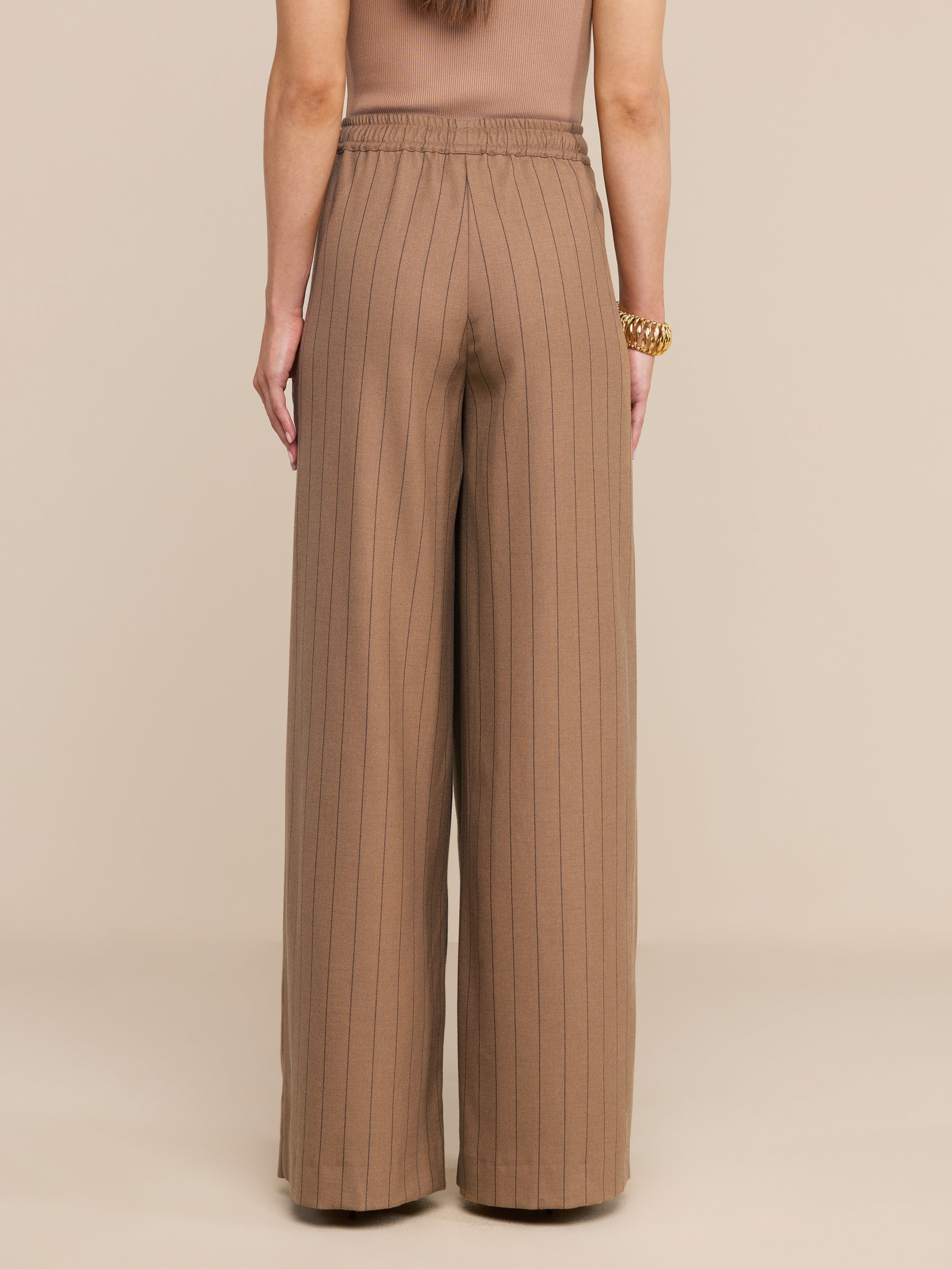 Une femme, de dos, porte un pantalon « Connie Drawstring » — un pantalon à taille haute et à jambes larges, à fines rayures — ainsi quun haut sans manches. Ses bras sont détendus le long du corps ; elle porte un gros bracelet en or au poignet droit. Larrière-plan est beige.