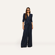 A woman stands against a plain light background, accessorized with sunglasses and long hair styled down, wearing the Connie Drawstring Trouser—part of a navy blue pinstripe suit paired with a fitted blazer—for a polished business casual look.