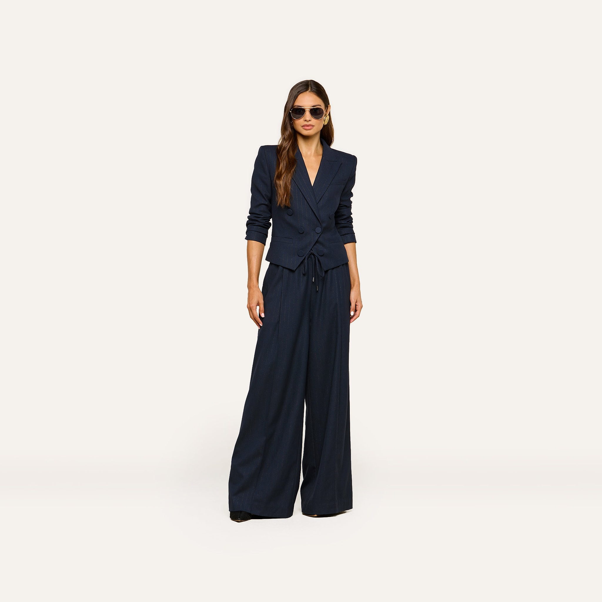 A woman stands against a plain light background, accessorized with sunglasses and long hair styled down, wearing the Connie Drawstring Trouser—part of a navy blue pinstripe suit paired with a fitted blazer—for a polished business casual look.
