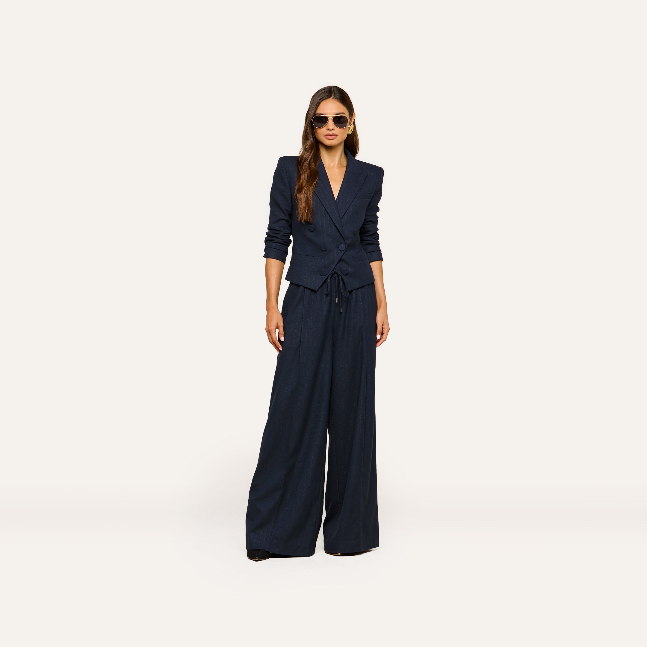 A woman stands against a plain light background, accessorized with sunglasses and long hair styled down, wearing the Connie Drawstring Trouser—part of a navy blue pinstripe suit paired with a fitted blazer—for a polished business casual look.
