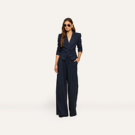 A woman wearing sunglasses and the Connie Drawstring Trouser in navy blue pinstripe, styled with matching wide-leg pants, stands with one hand in her pocket against a plain, light background.