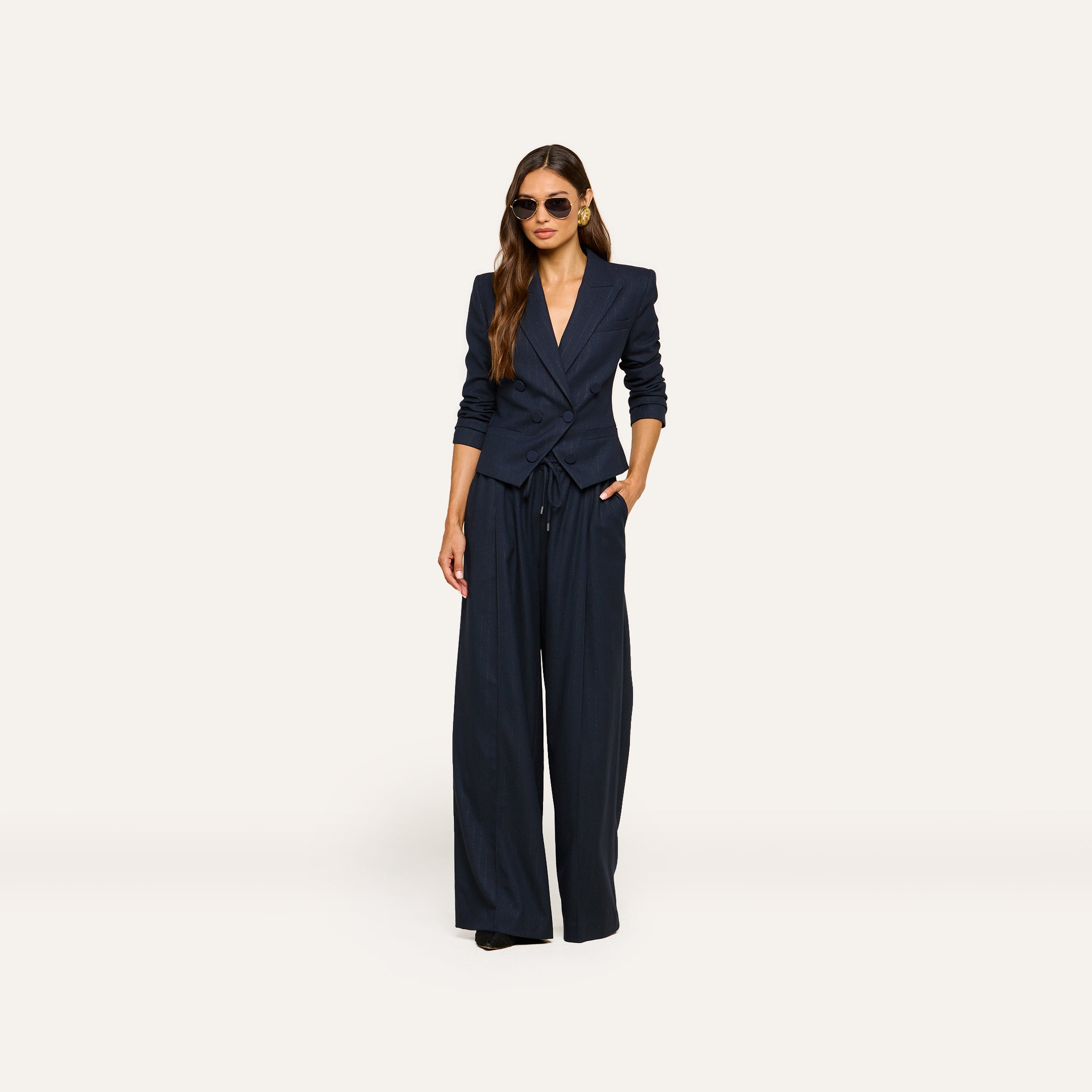 A woman wearing sunglasses and the Connie Drawstring Trouser in navy blue pinstripe, styled with matching wide-leg pants, stands with one hand in her pocket against a plain, light background.