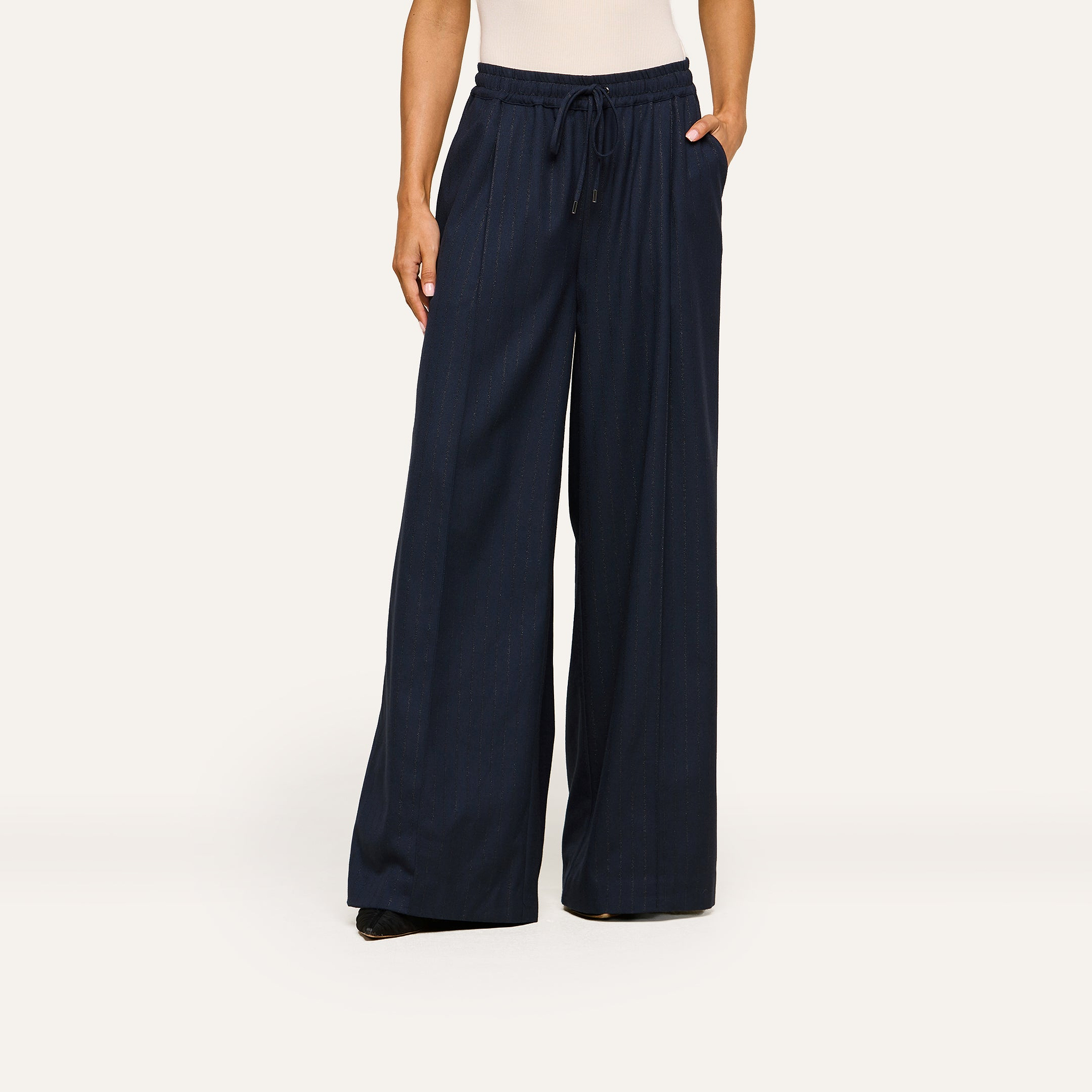 A person stands against a plain background wearing the Connie Drawstring Trouser—a high-waisted, wide-leg navy blue pant with a drawstring waist and side pockets—styled in a business casual look and paired with a sleeveless beige top.