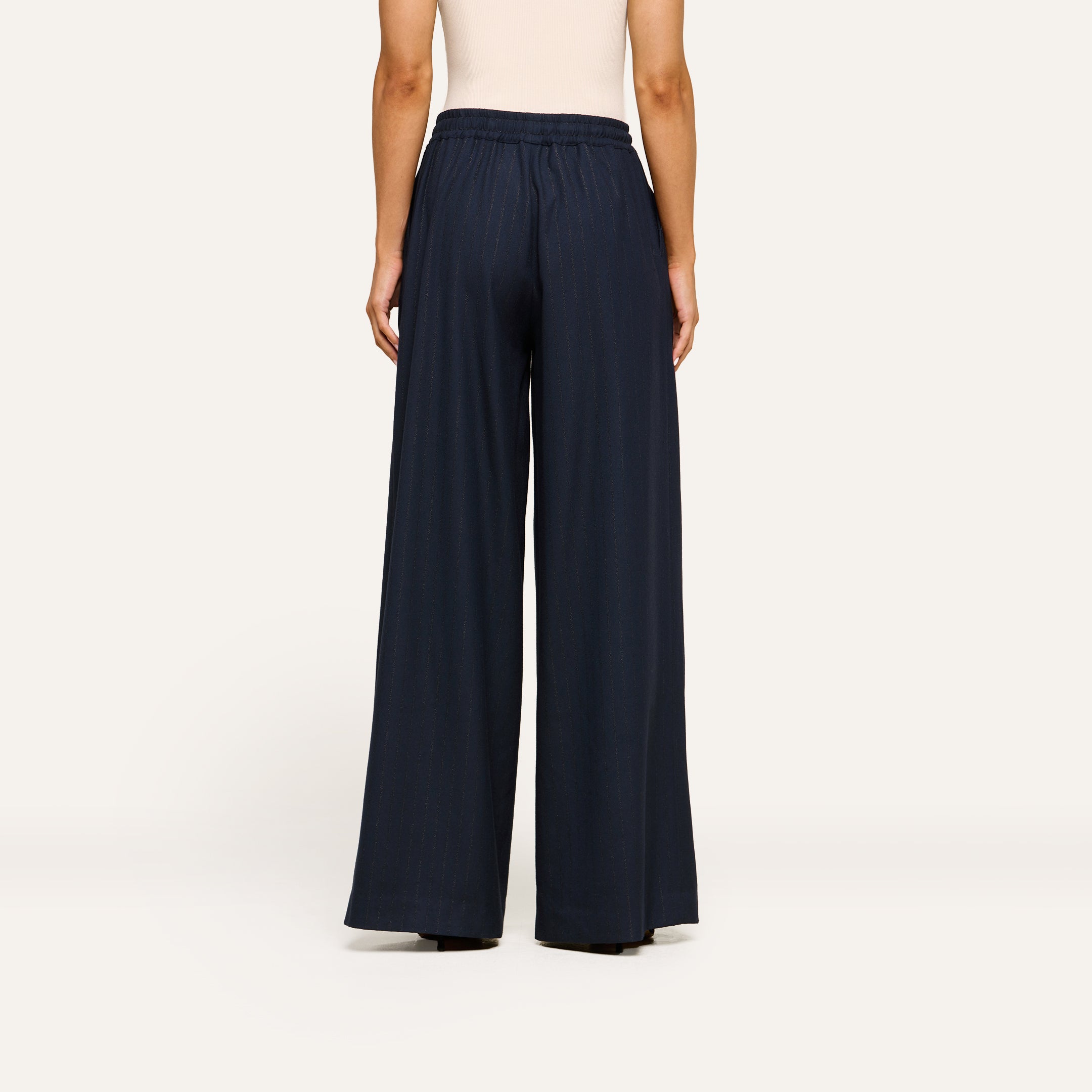 A person stands facing away, wearing a sleeveless beige top paired with the Connie Drawstring Trouser in navy blue—a refined and modern ensemble that captures a business casual style against a plain, light-colored background.