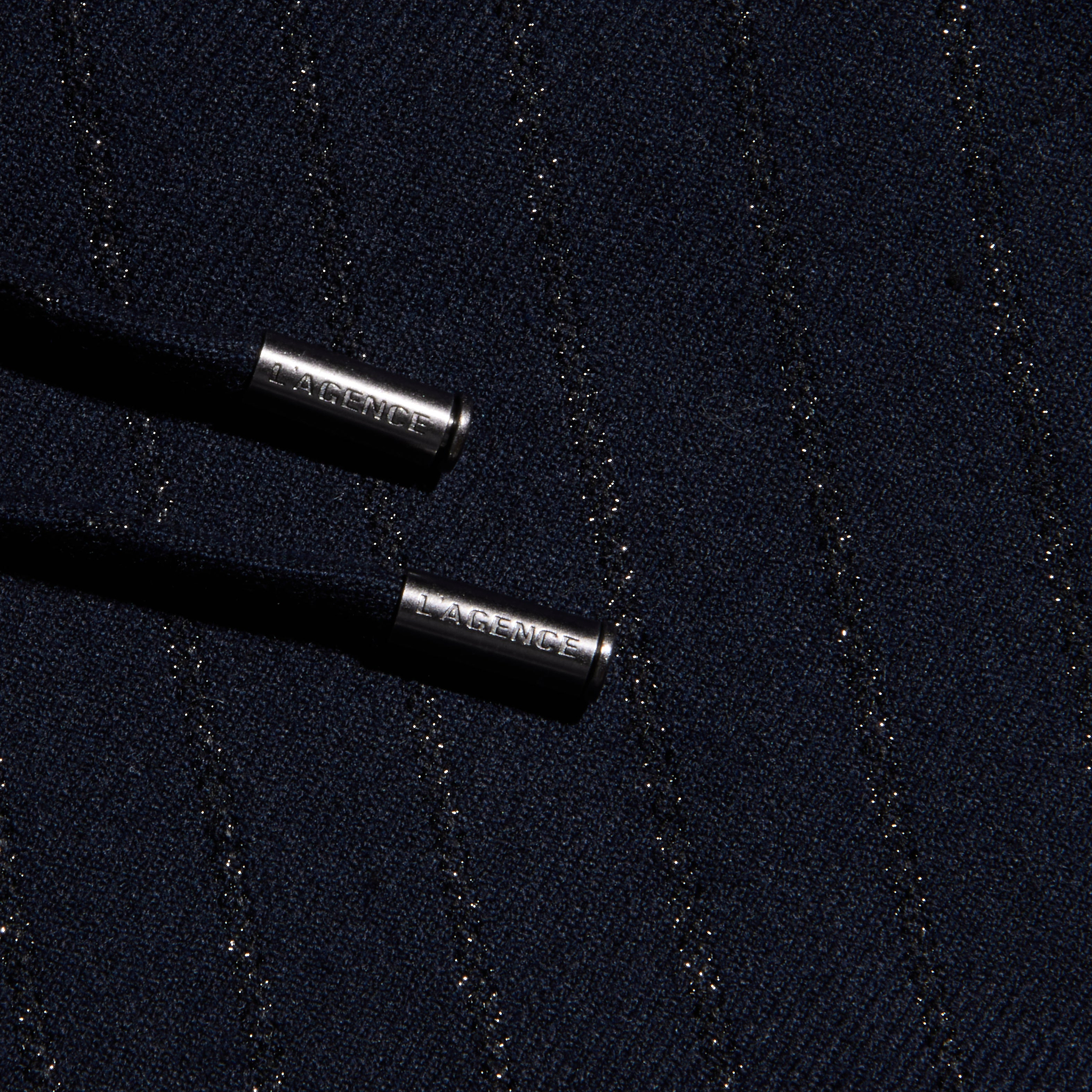 Close-up of the Connie Drawstring Trouser’s dark textured fabric, woven with diagonal metallic threads for a pinstripe effect, and featuring two black drawstrings finished with silver metal tips engraved with LAGENCE.