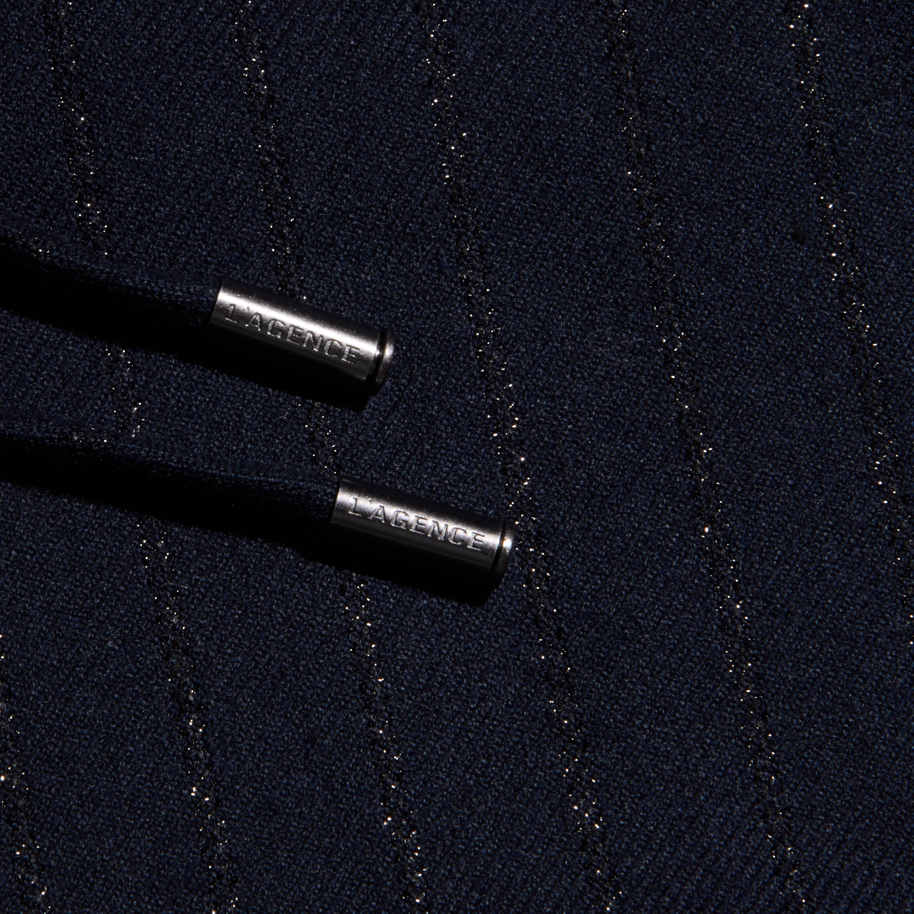 Close-up of the Connie Drawstring Trouser’s dark textured fabric, woven with diagonal metallic threads for a pinstripe effect, and featuring two black drawstrings finished with silver metal tips engraved with LAGENCE.