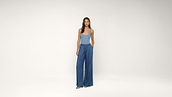 A woman stands against a plain white background wearing a light blue tank top, wide-leg blue pants, and sandals, with her hands in her pockets.