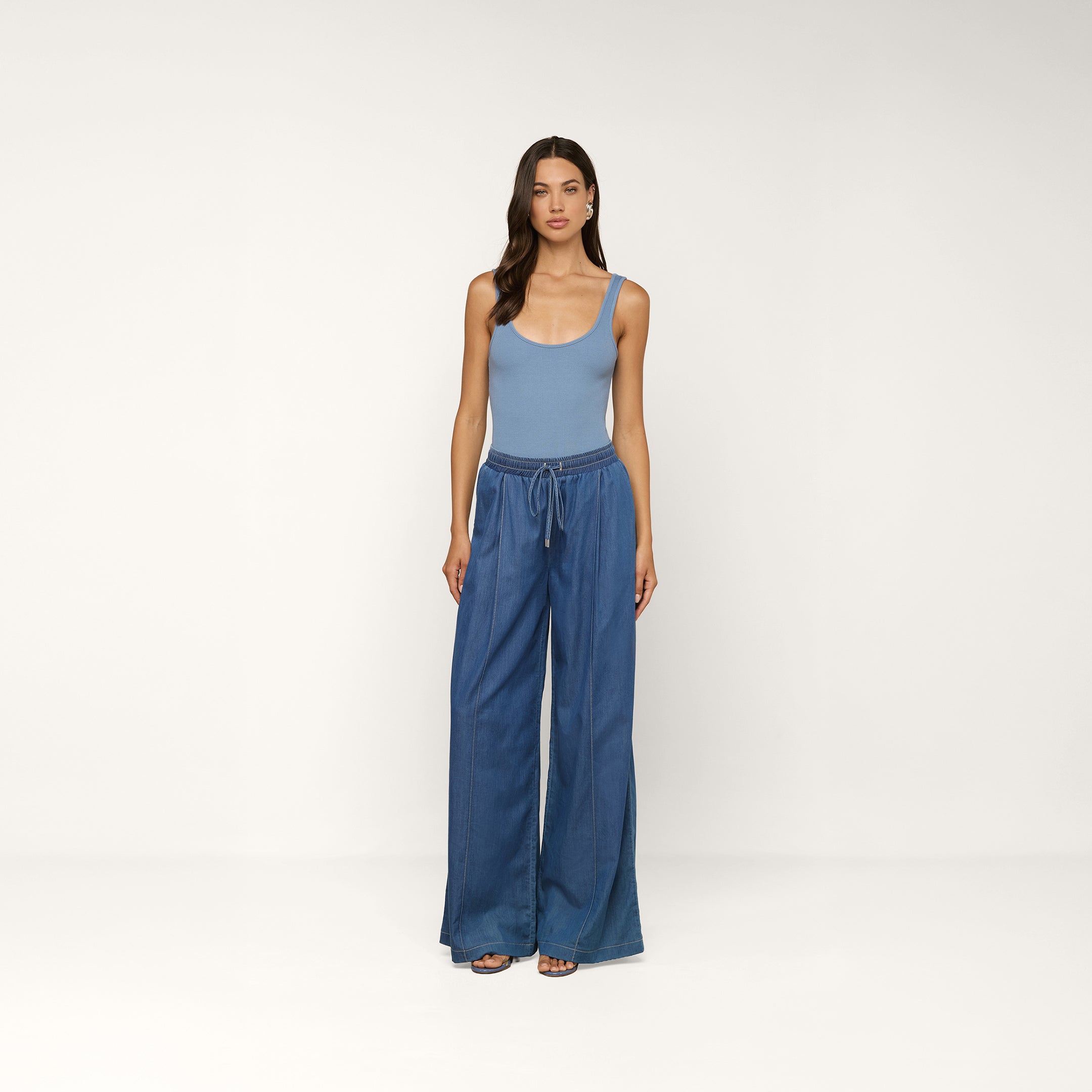 Vor einem beigen Hintergrund steht eine Frau, die ein hellblaues, ärmelloses Tanktop und die „Connie Drawstring Jean“ trägt – eine Jeans aus federleichtem Denim mit lockerer Passform und Kordelzug in der Taille. Dazu trägt sie Sandalen mit offener Zehenpartie und hat langes, welliges Haar.