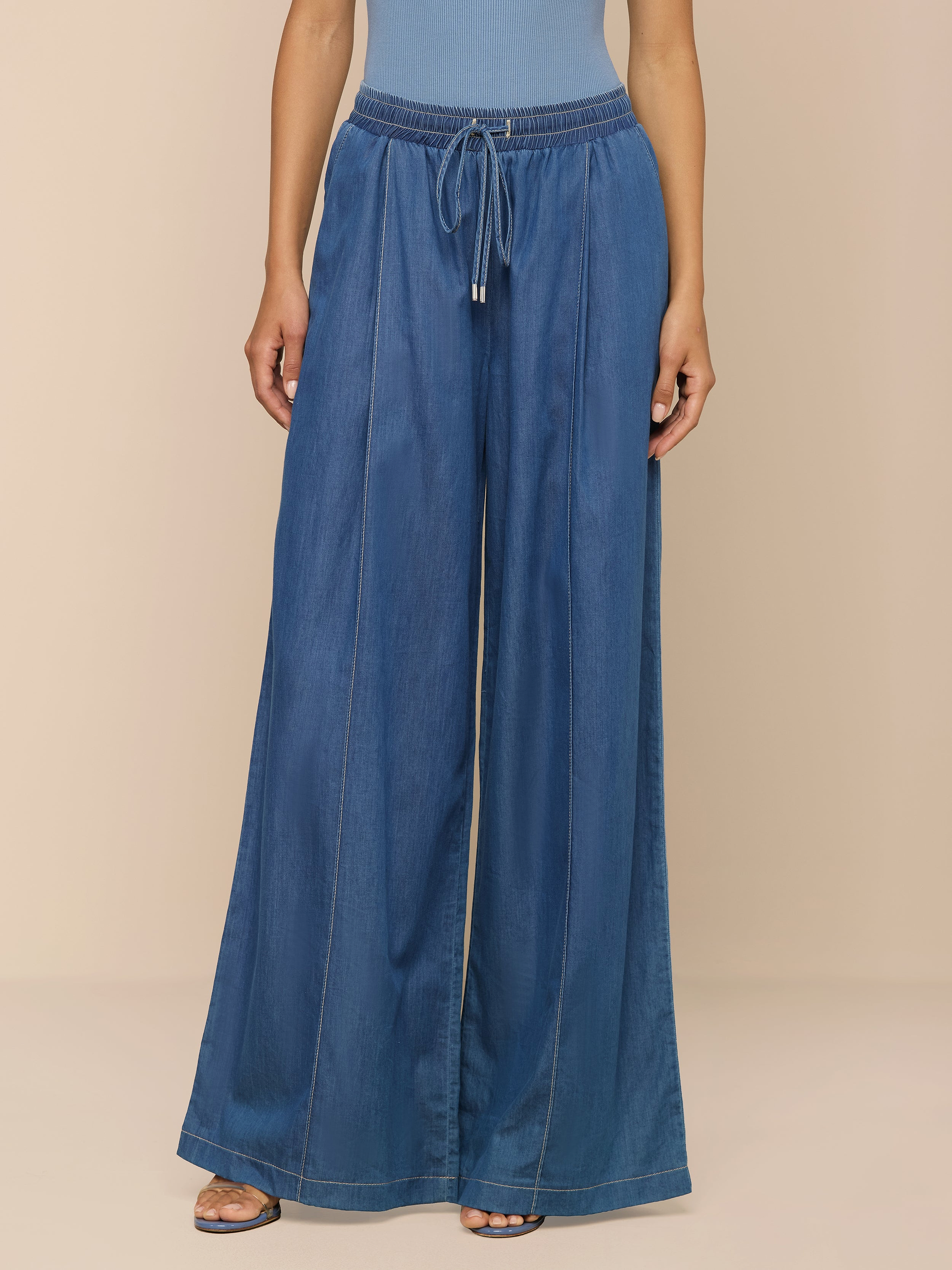 Eine Person, die die „Connie Drawstring Jean“ trägt – lockere Passform, federleichter Denim mit Kordelzug in der Taille und sichtbaren Nahtdetails –, kombiniert mit Sandalen mit offener Zehenpartie und einem blauen Oberteil, steht vor einem beigen Hintergrund.