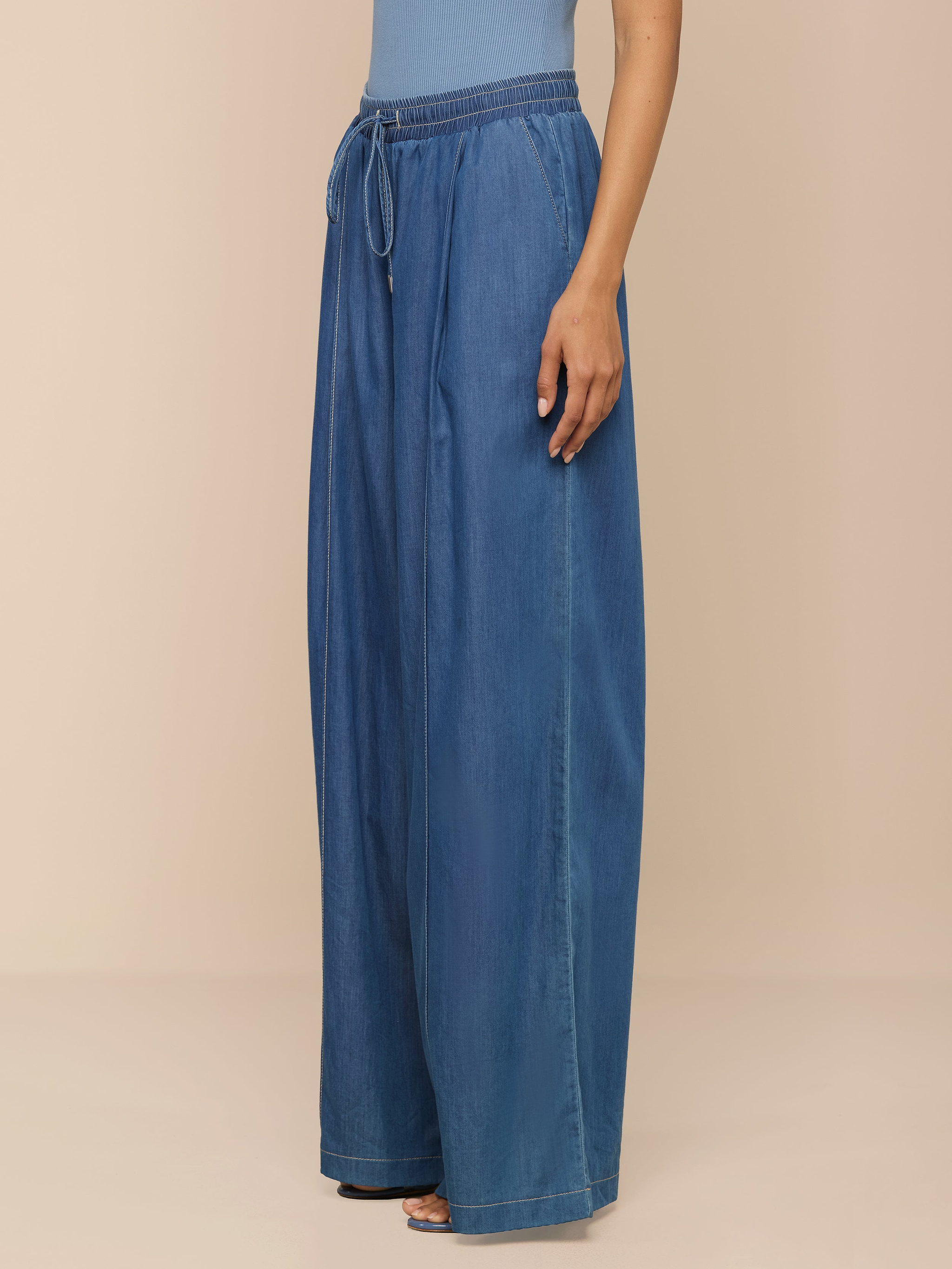 Eine Person steht vor einem neutralen Hintergrund und trägt die „Connie Drawstring Jean“ – eine federleichte Hose mit weitem Bein, hoher Taille, Kordelzug und Seitentaschen –, kombiniert mit einem hellblauen Oberteil.