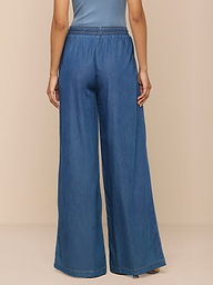 Vor einem neutralen beigen Hintergrund ist eine Person von hinten zu sehen, die die „Connie Drawstring Jean“ trägt – eine blaue Hose mit weitem Bein und lockerer Passform aus federleichtem Denim und elastischem Bund –, kombiniert mit einem blauen Oberteil.