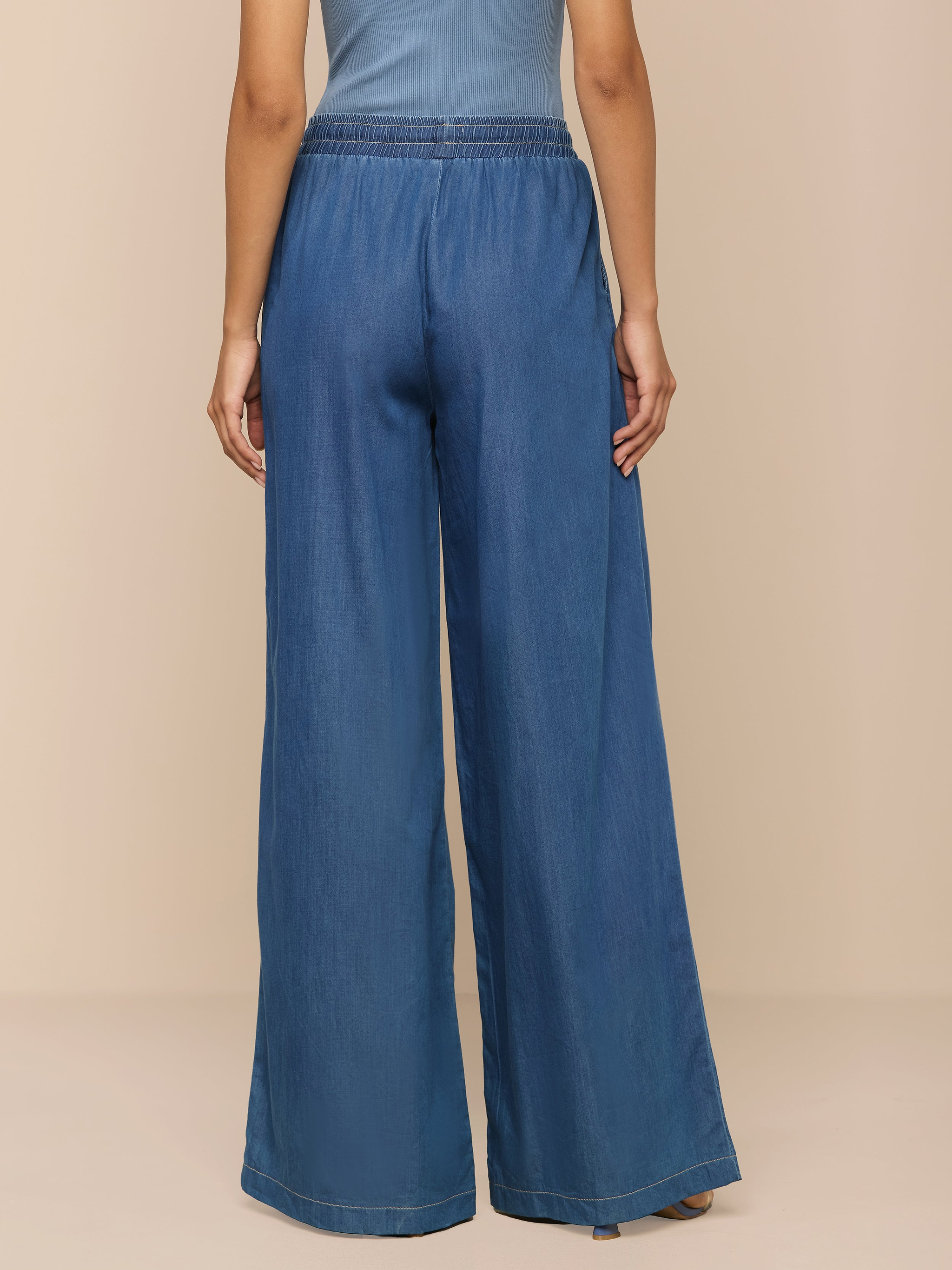 Vor einem neutralen beigen Hintergrund ist eine Person von hinten zu sehen, die die „Connie Drawstring Jean“ trägt – eine blaue Hose mit weitem Bein und lockerer Passform aus federleichtem Denim und elastischem Bund –, kombiniert mit einem blauen Oberteil.
