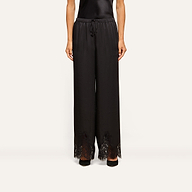 A person is wearing the Mirel Satin Lace Pant, featuring a black drawstring waist and lace trim at the hem, paired with black shoes. Only the lower half of the body is visible against a plain background.