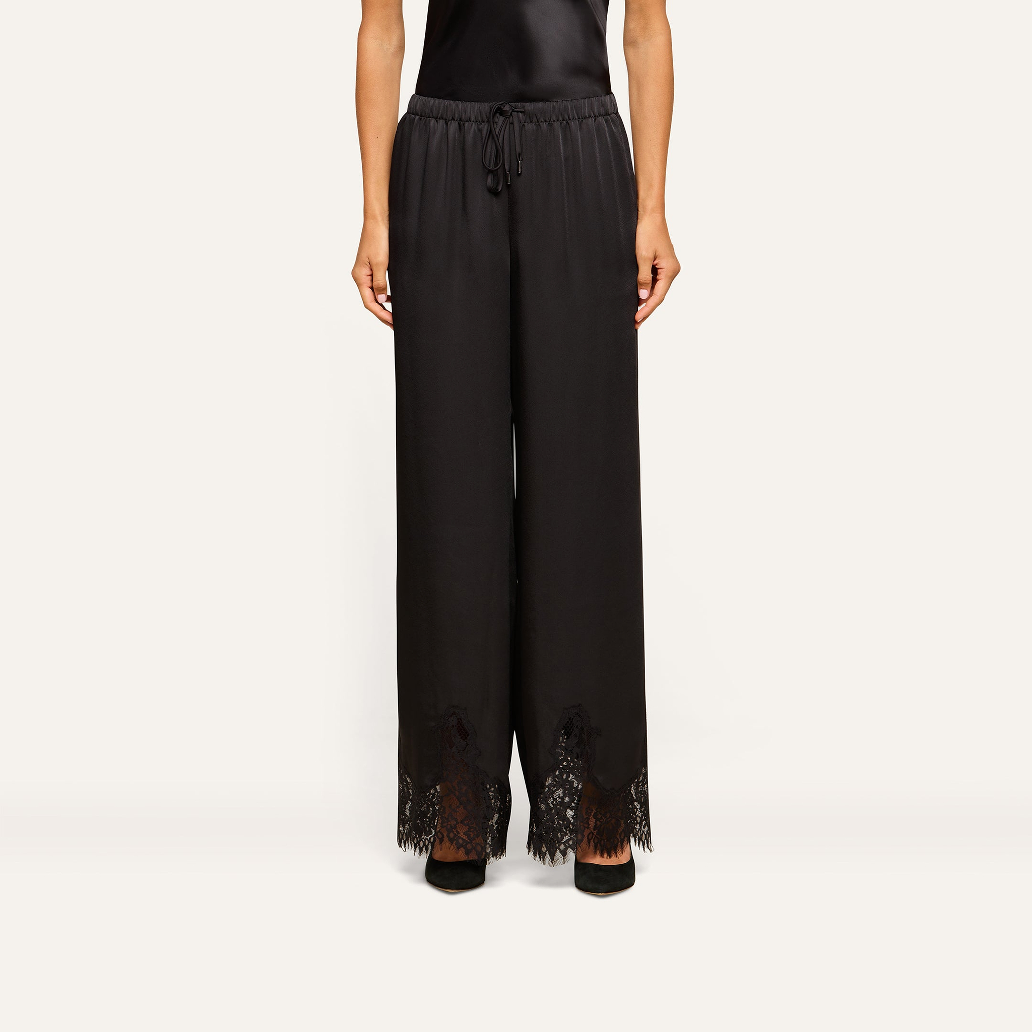 A person is wearing the Mirel Satin Lace Pant, featuring a black drawstring waist and lace trim at the hem, paired with black shoes. Only the lower half of the body is visible against a plain background.