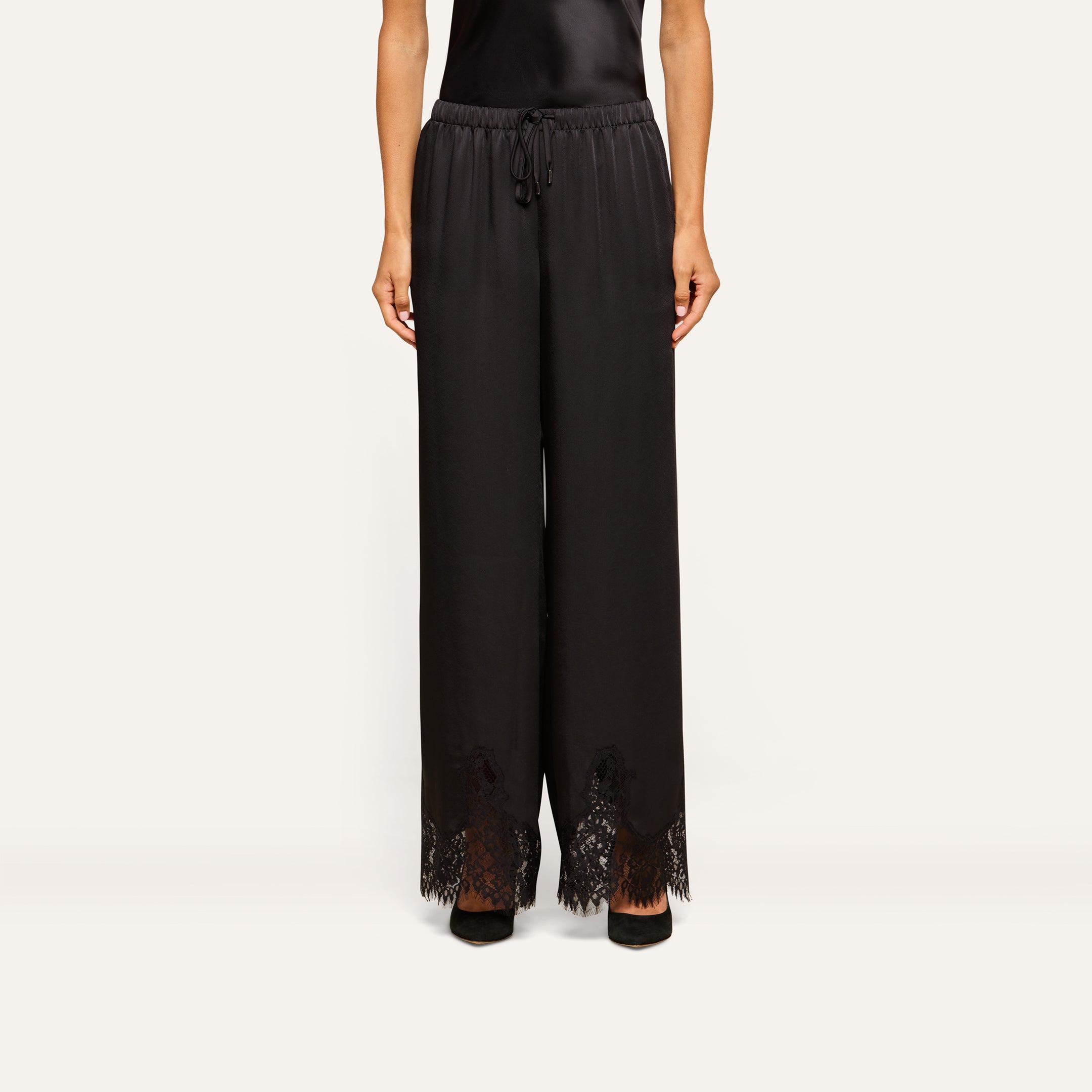 A person is wearing the Mirel Satin Lace Pant, featuring a black drawstring waist and lace trim at the hem, paired with black shoes. Only the lower half of the body is visible against a plain background.