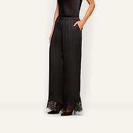 Wearing the Mirel Satin Lace Pant, which features a loose fit and delicate lace trim at the hem, a woman pairs it with a sleeveless black top and black pointed heels as she stands against a plain light background.