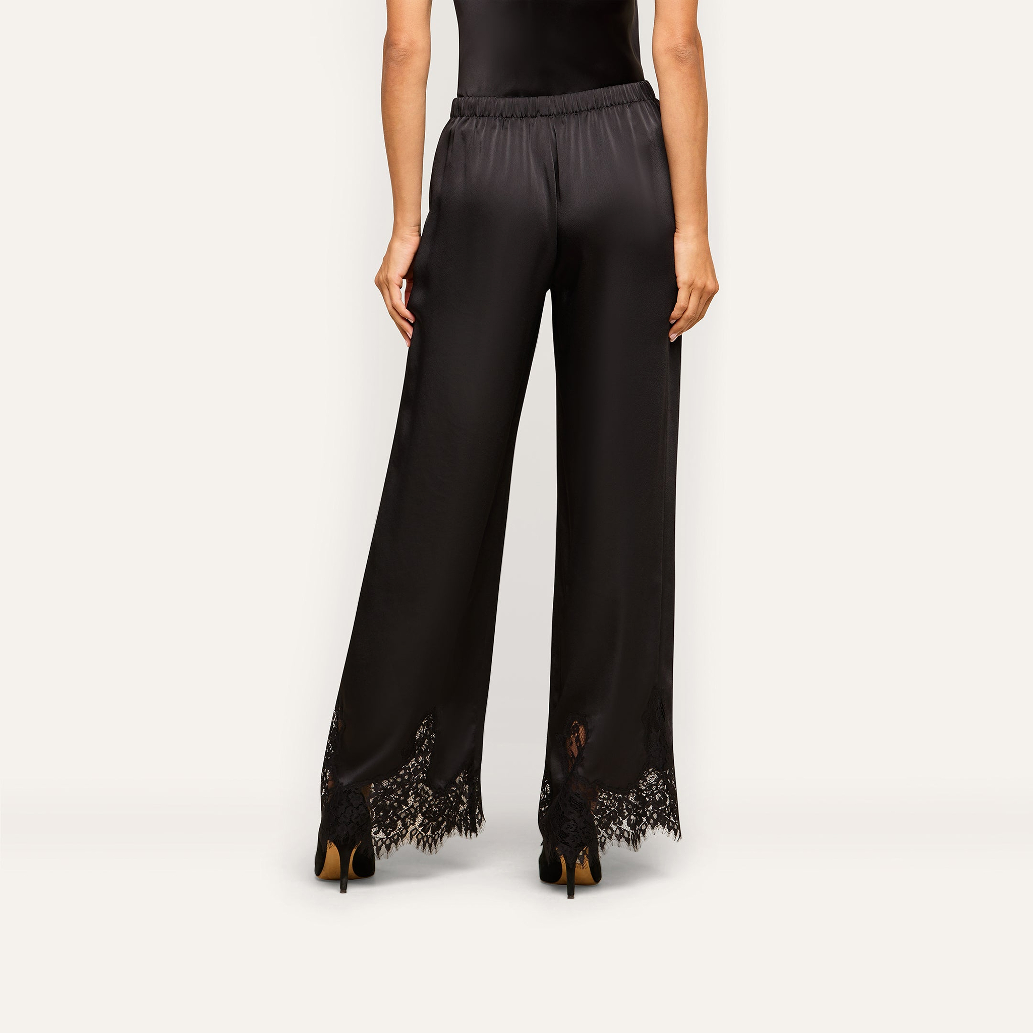 A woman, seen from behind, models the Mirel Satin Lace Pant—featuring black satin fabric, wide legs, and delicate lace trim at the hem—against a plain light background.