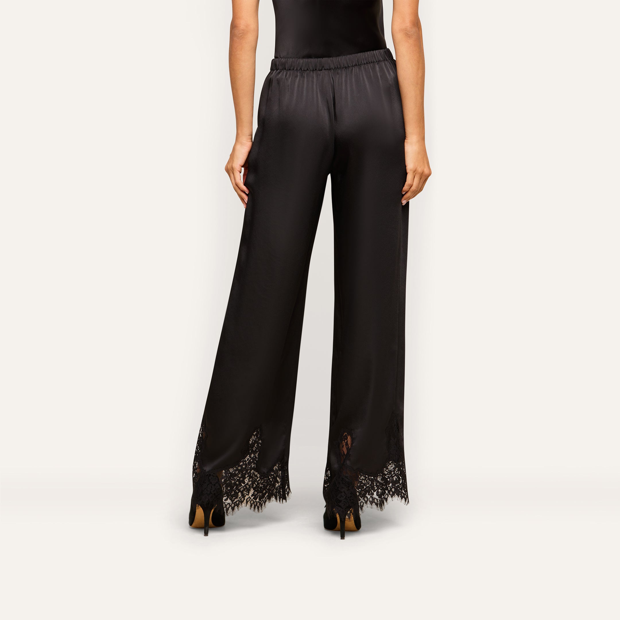 A woman, seen from behind, models the Mirel Satin Lace Pant—featuring black satin fabric, wide legs, and delicate lace trim at the hem—against a plain light background.