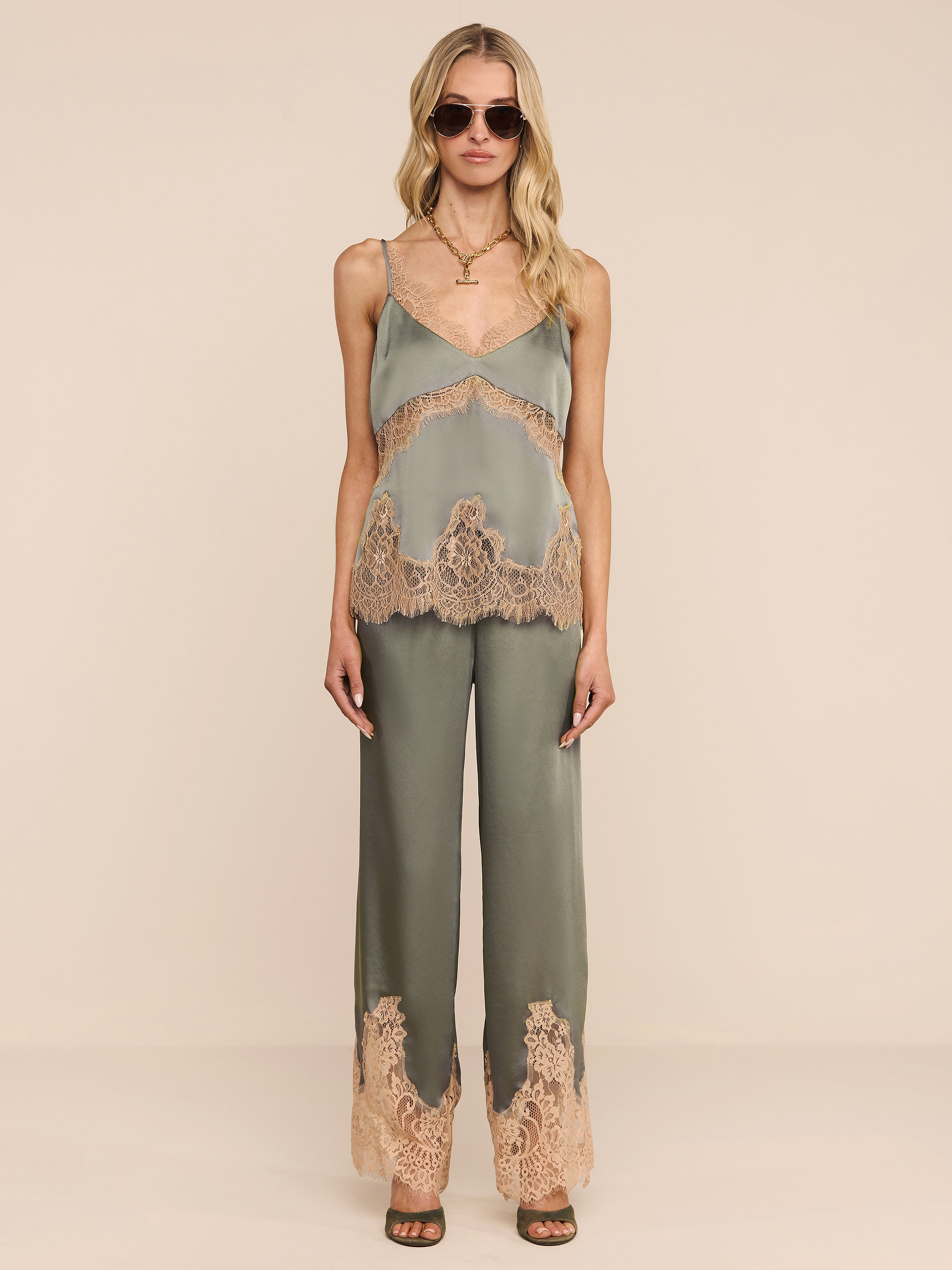 A woman stands against a plain white background wearing the Mirel Pant in olive green, featuring lace trim and a drawstring waist. She pairs it with a matching camisole, sunglasses, open-toe heels, and her long blonde hair.