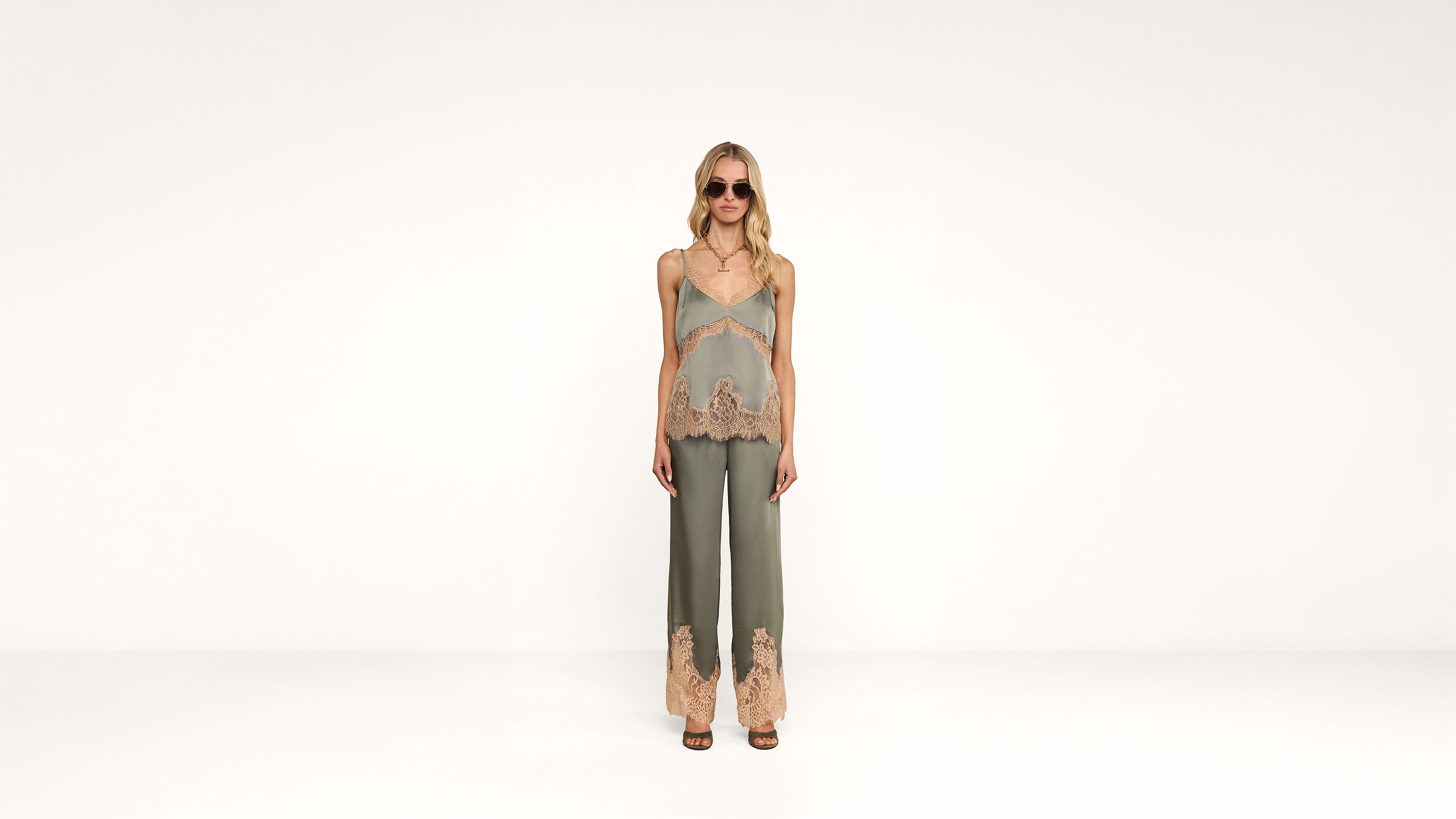 A woman wearing sunglasses, a green camisole top, and matching green pants with beige lace trim stands against a plain white background.