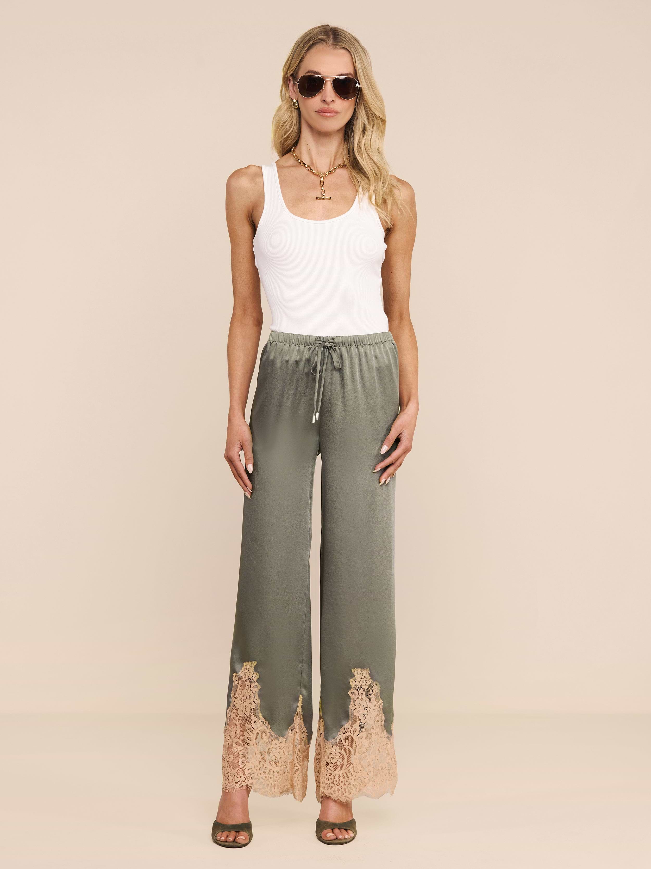 A woman stands facing forward against a plain white background, wearing sunglasses, a white tank top, and the Mirel Pant—green satin pants featuring beige lace trim and a drawstring waist.
