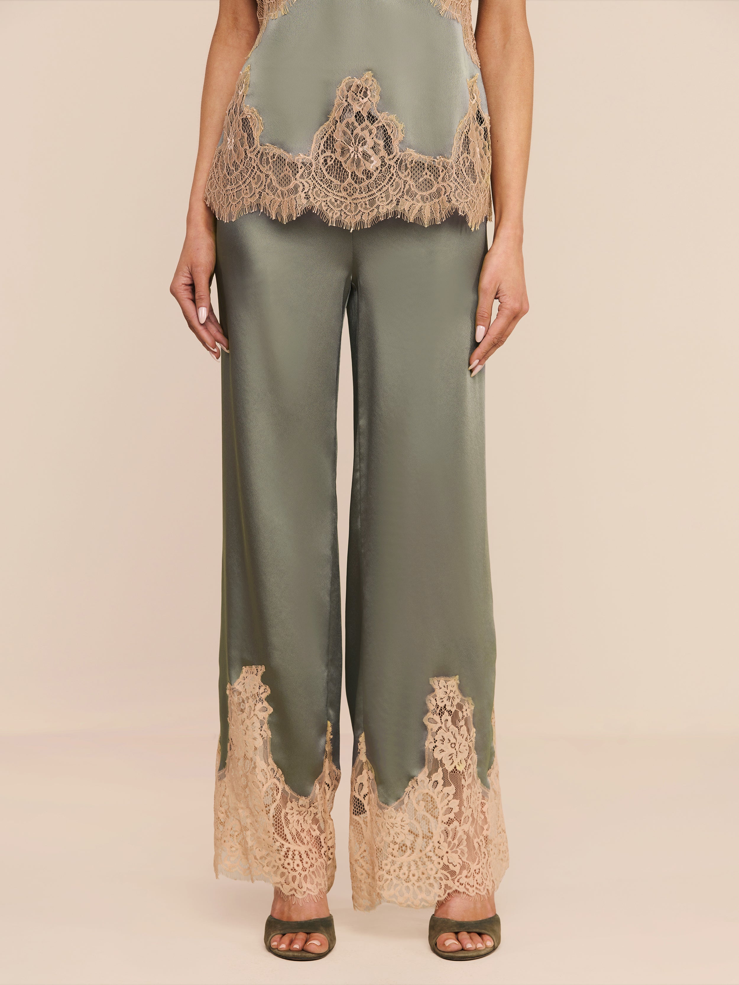 Wearing the Mirel Pant in olive green satin, featuring a drawstring waist and lace trim at the hem, paired with a coordinating top adorned with matching lace details, the individual is posed against a plain white backdrop.