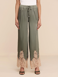Wearing the Mirel Pant in olive green—with a wide-leg silhouette, lace trim, and a drawstring waist—a person pairs it with a white top and open-toed sandals, standing against a plain white background.