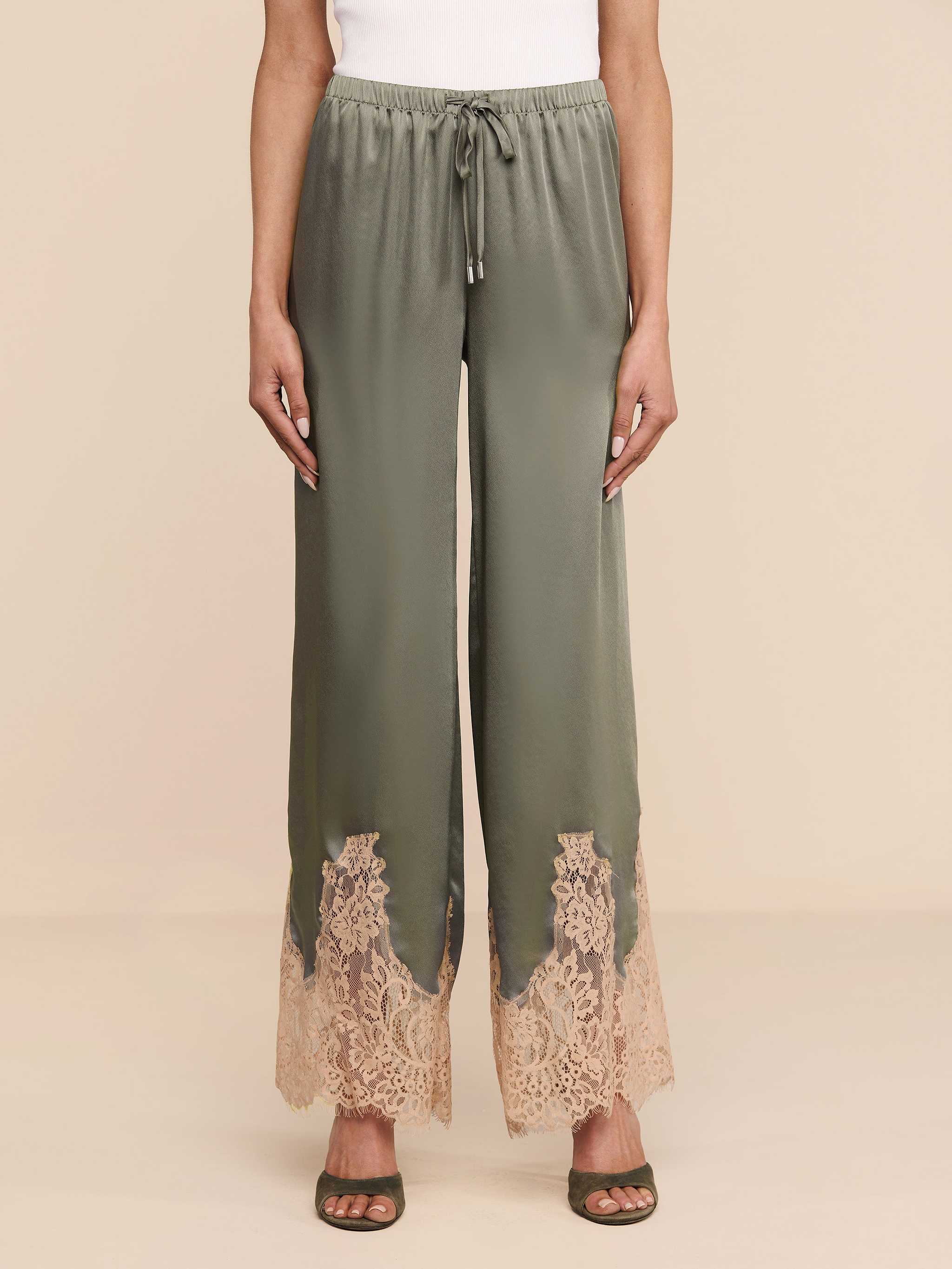 Wearing the Mirel Pant in olive green—with a wide-leg silhouette, lace trim, and a drawstring waist—a person pairs it with a white top and open-toed sandals, standing against a plain white background.