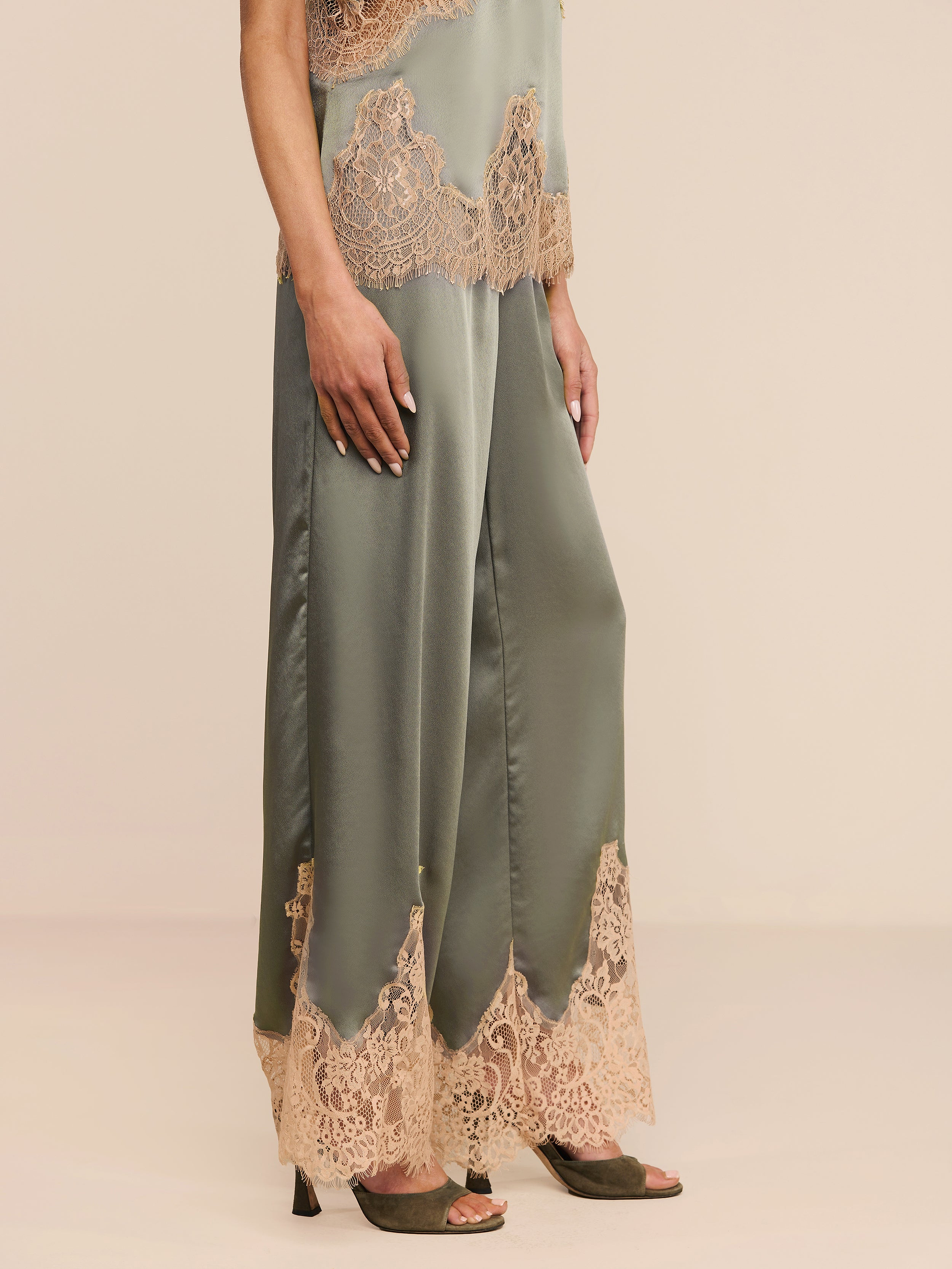 A person wearing the Mirel Pant—olive satin pants featuring a drawstring waist and beige lace trim at the hem—paired with a matching lace-trimmed top and olive open-toe high heels, stands against a plain white background.
