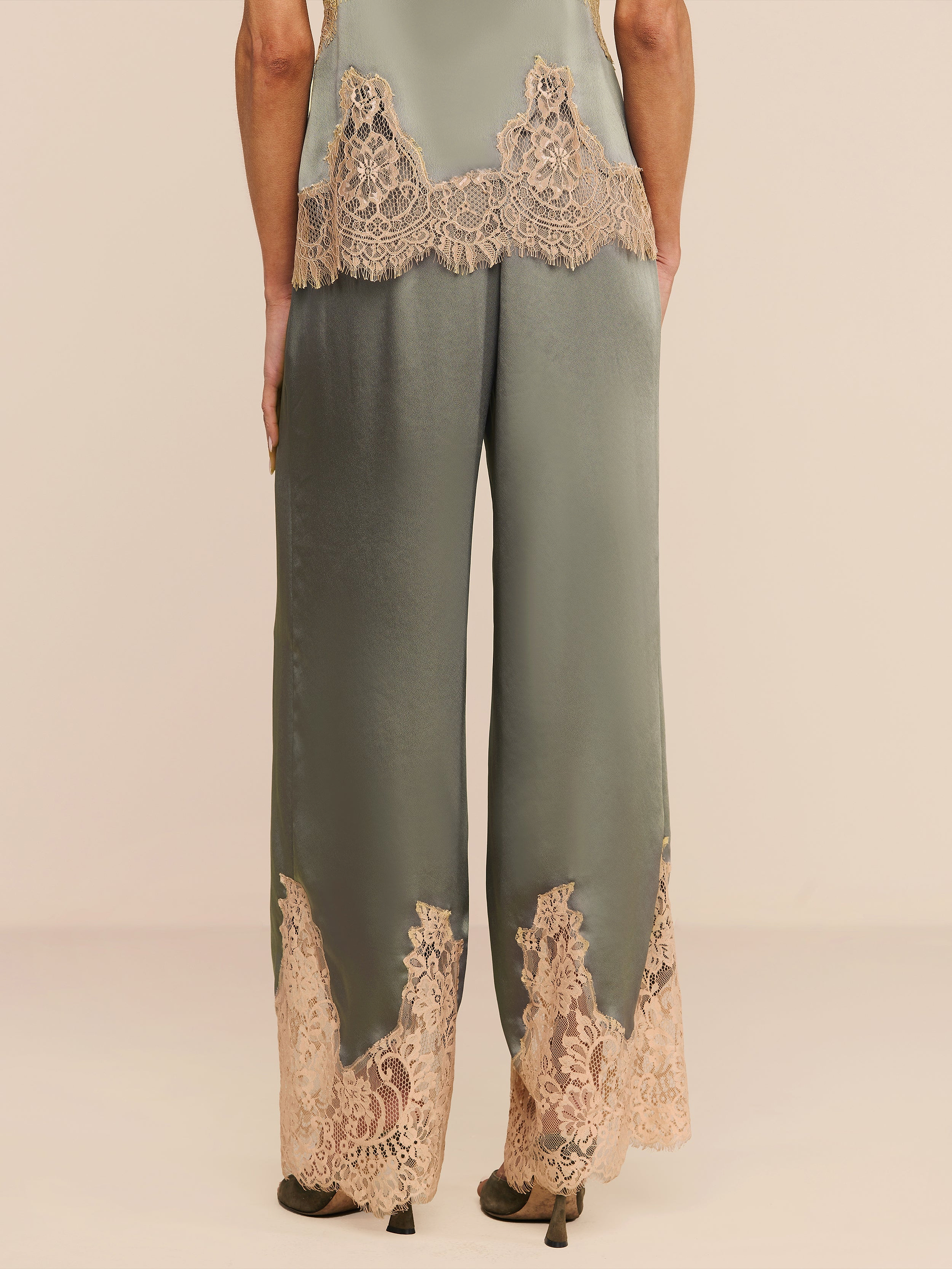 A person stands facing away, wearing the Mirel Pant in silky olive green satin featuring a drawstring waist and beige lace trim at the hem and waist, paired with matching high heels against a plain white background.