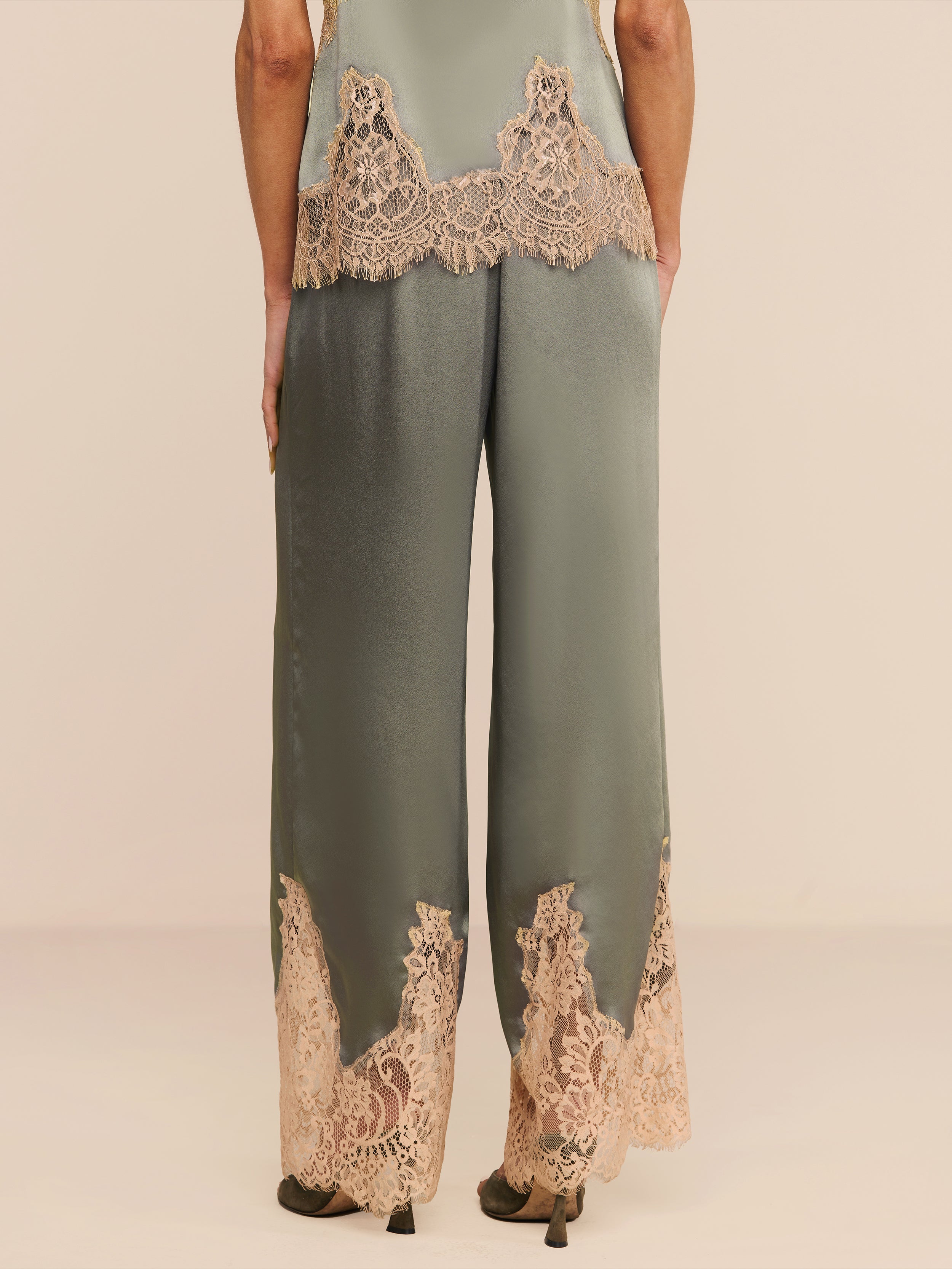 A person stands facing away, wearing the Mirel Pant in silky olive green satin featuring a drawstring waist and beige lace trim at the hem and waist, paired with matching high heels against a plain white background.
