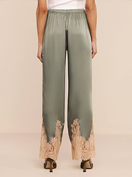 Model is seen from behind wearing the Mirel Pant in olive green satin, featuring a drawstring waist, beige lace trim at the hem, and a relaxed fit, styled with heeled shoes.
