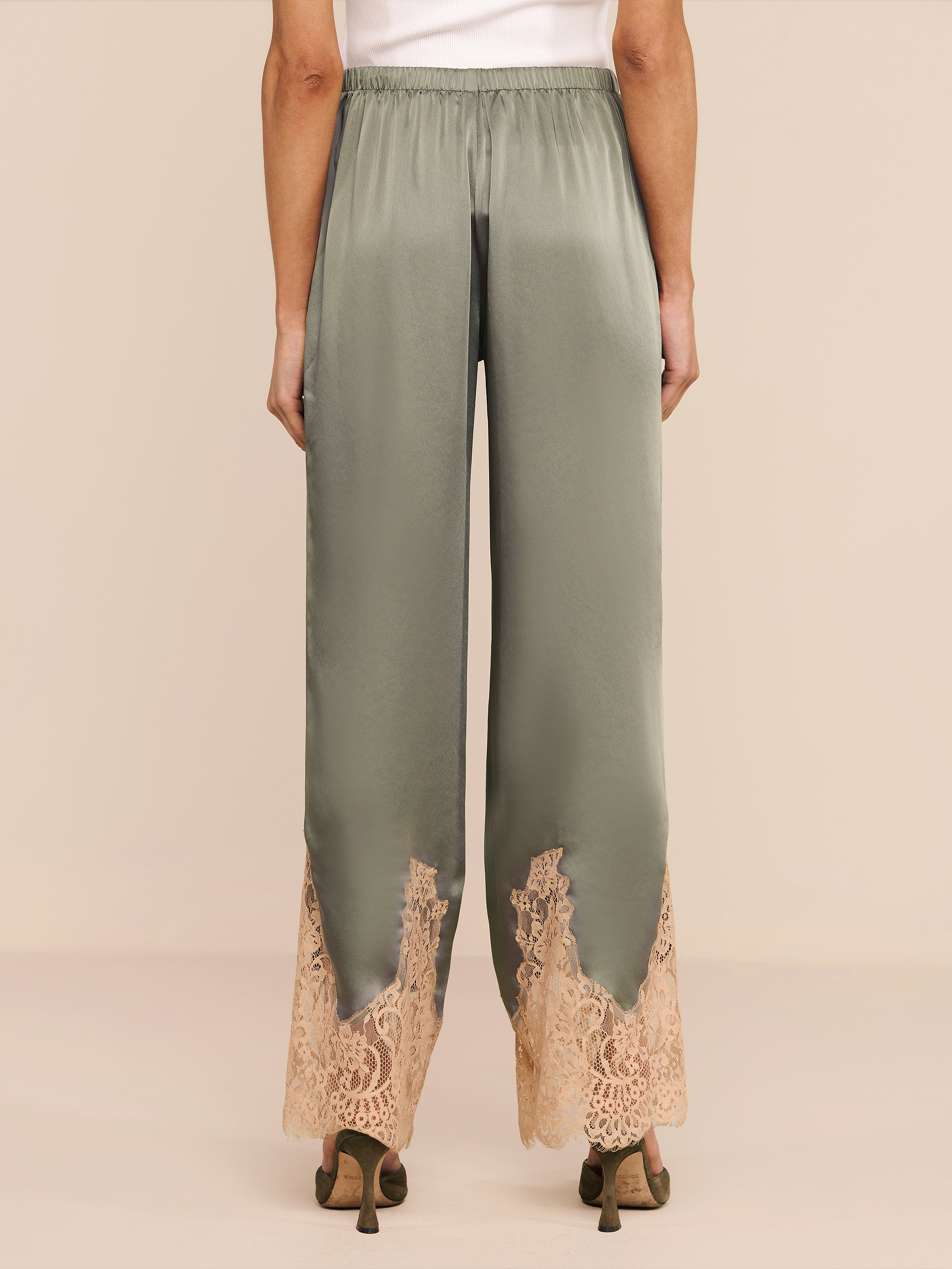 Model is seen from behind wearing the Mirel Pant in olive green satin, featuring a drawstring waist, beige lace trim at the hem, and a relaxed fit, styled with heeled shoes.