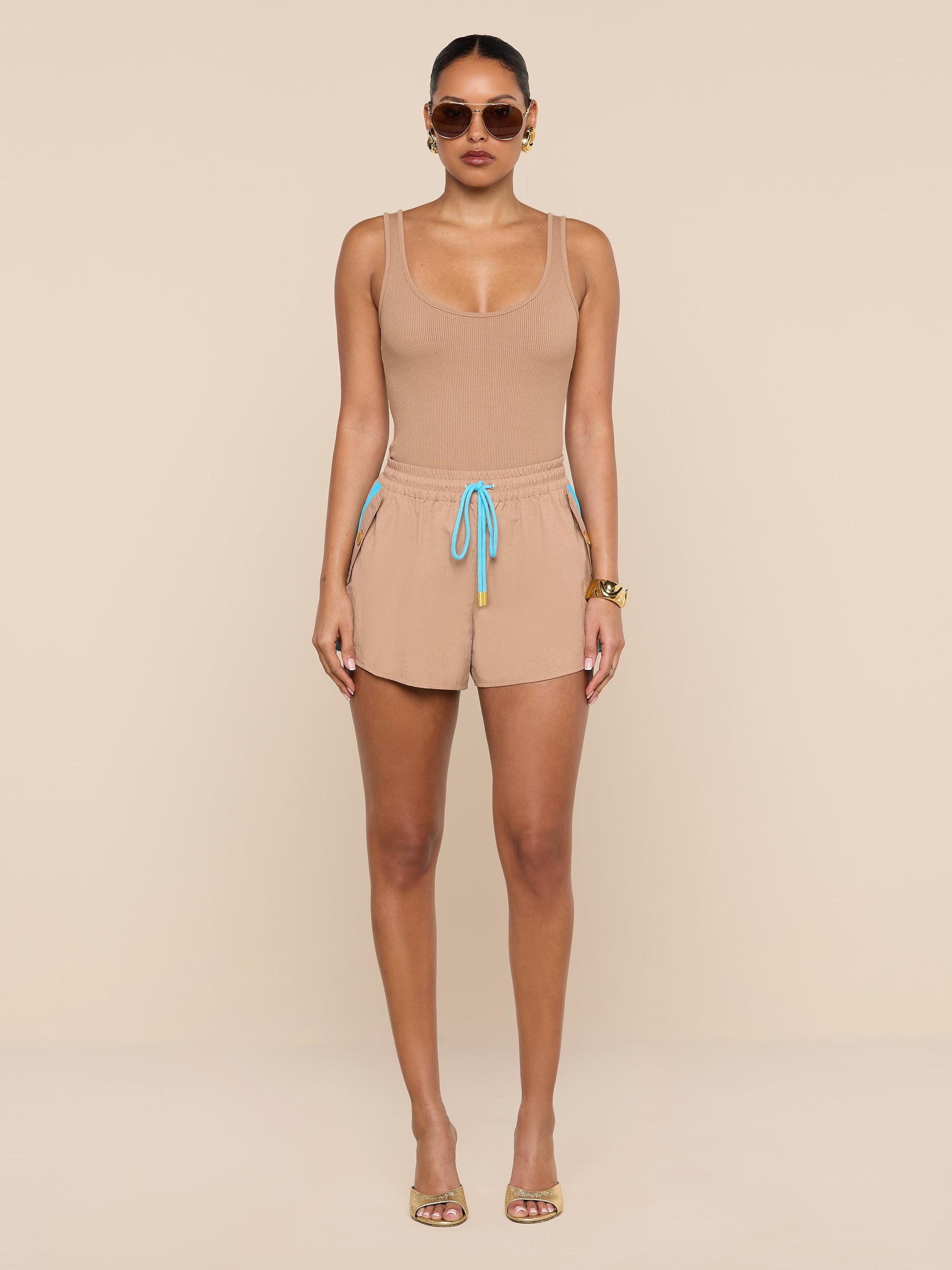 A woman stands against a white background, wearing a beige crepe nylon tank top and matching Grayson Nylon Shorts with a blue drawstring, paired with gold sandals, sunglasses, and gold bracelets.