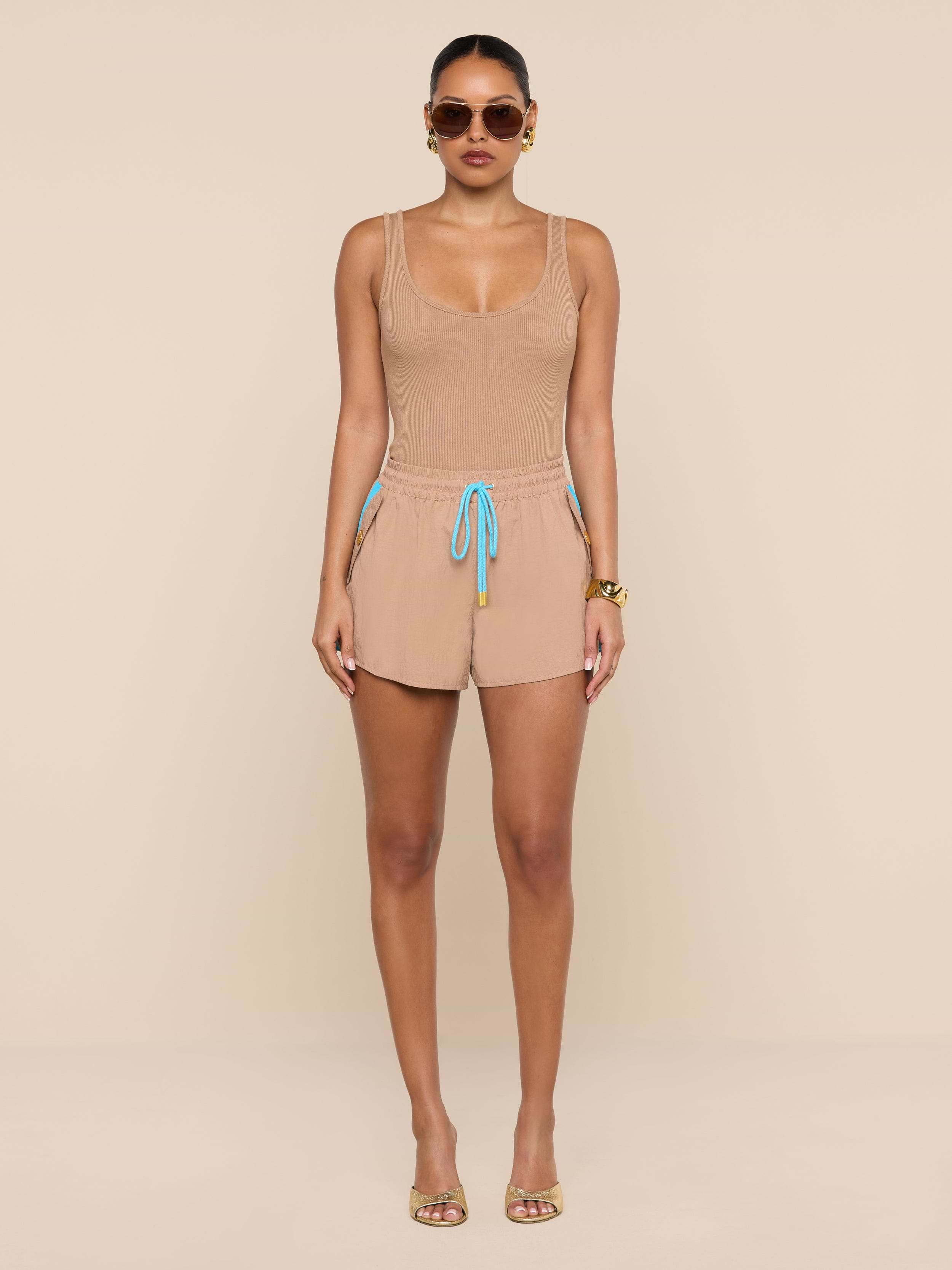 A woman stands against a white background, wearing a beige crepe nylon tank top and matching Grayson Nylon Shorts with a blue drawstring, paired with gold sandals, sunglasses, and gold bracelets.
