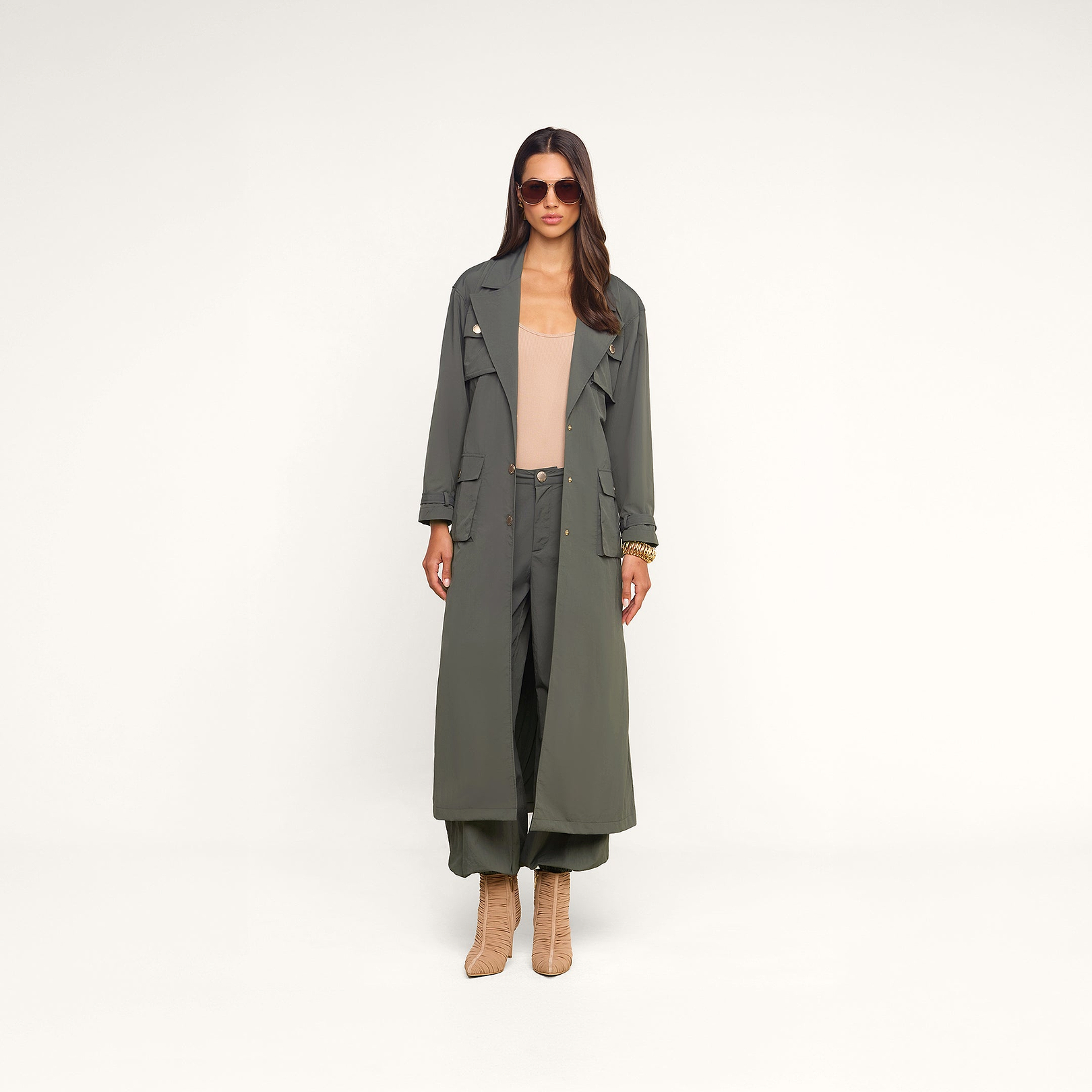 A woman wearing sunglasses, a long ash olive green trench coat with cargo pockets, the Greer Nylon Jogger in olive, a beige top, and tan heeled boots stands against a plain white background.