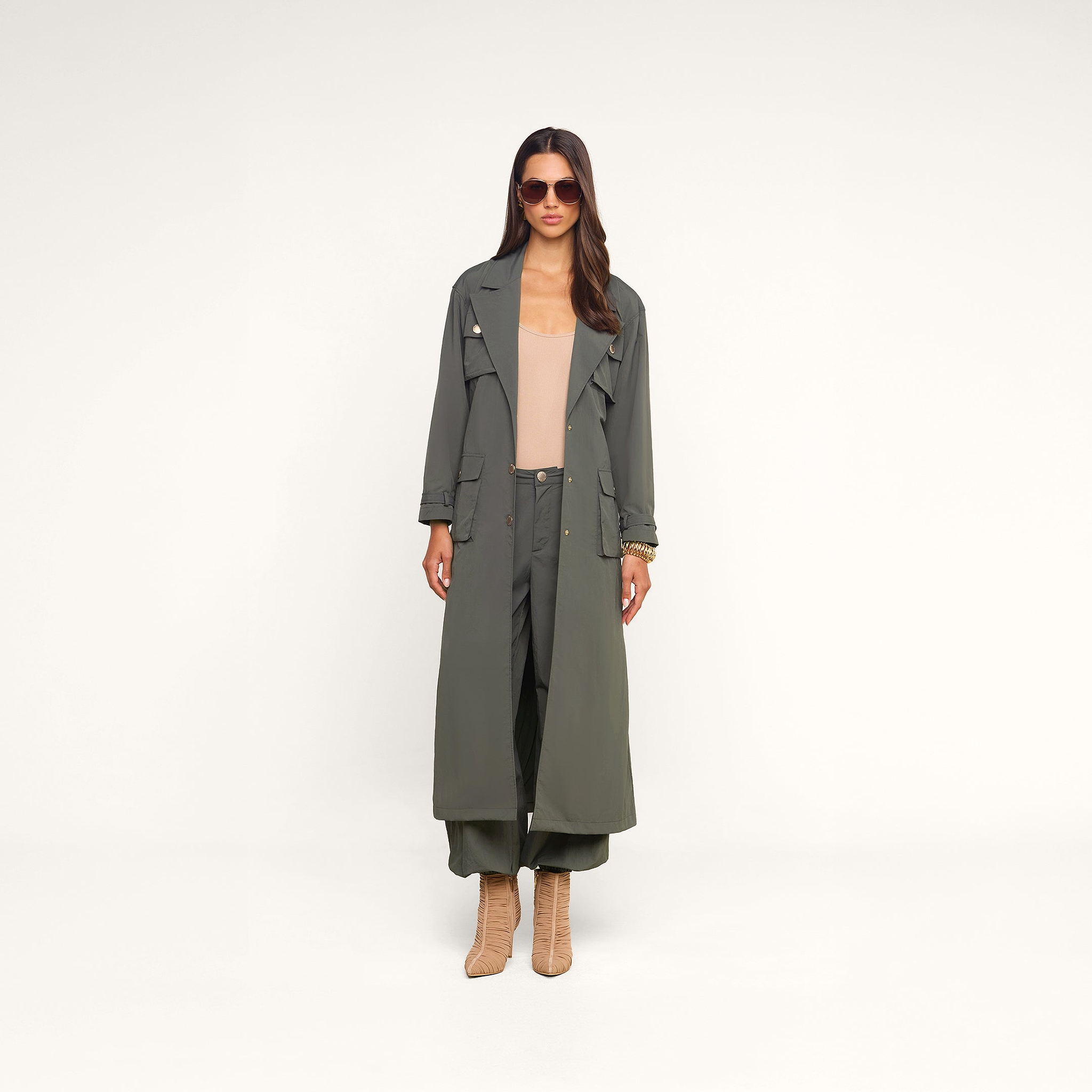 Woman wearing a long ash olive green trench coat with cargo pockets, Greer Nylon Jogger pants in olive, a tan top, heeled tan boots, sunglasses, and gold bracelets, standing against a beige background.