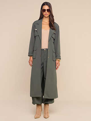 Woman wearing a long ash olive green trench coat with cargo pockets, Greer Nylon Jogger pants in olive, a tan top, heeled tan boots, sunglasses, and gold bracelets, standing against a beige background.