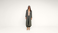 A woman stands facing forward, wearing a long olive green coat, matching wide-leg pants, a beige top, tan heeled boots, and sunglasses, against a plain white background.