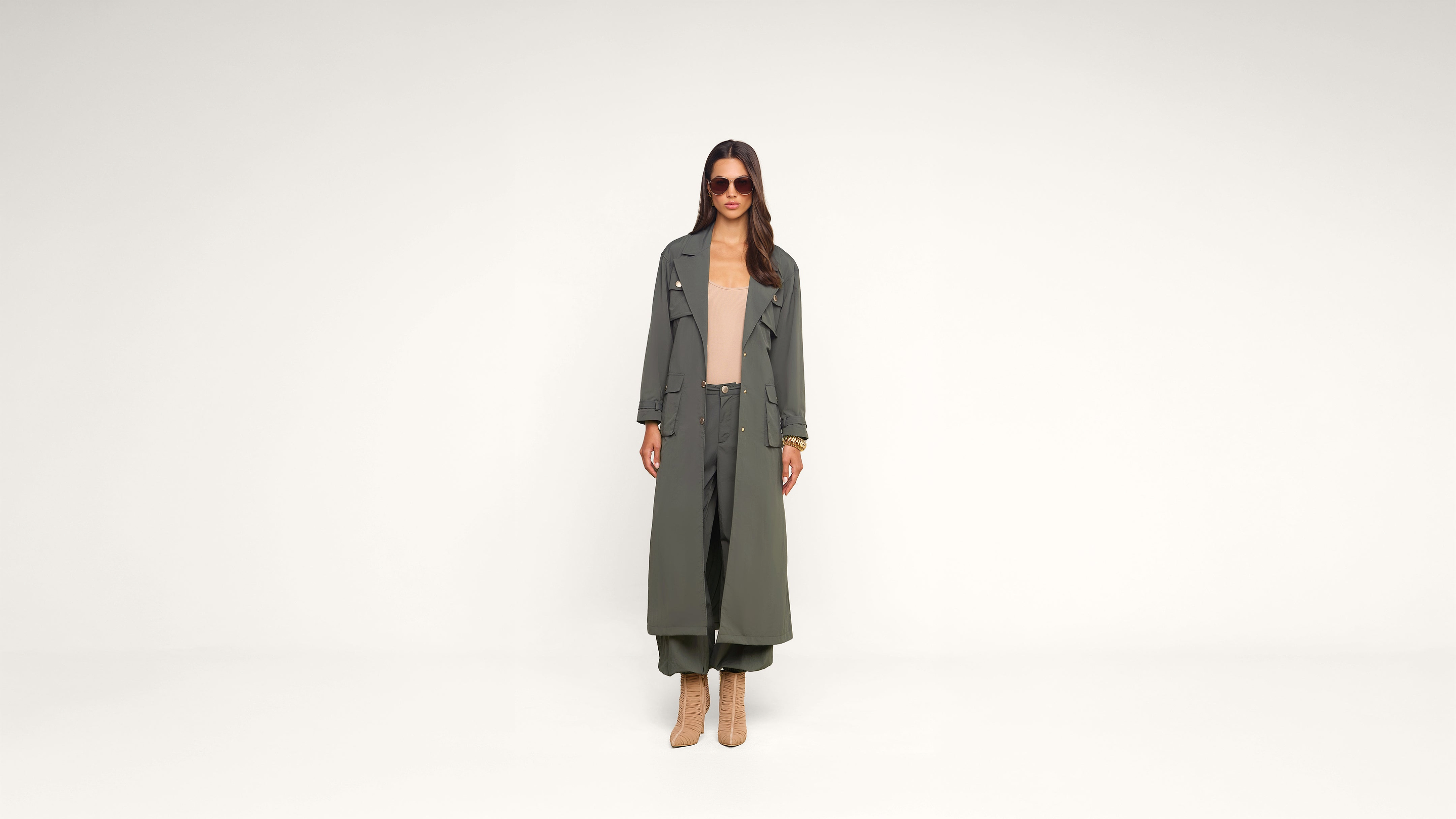 A woman stands facing forward, wearing a long olive green coat, matching wide-leg pants, a beige top, tan heeled boots, and sunglasses, against a plain white background.