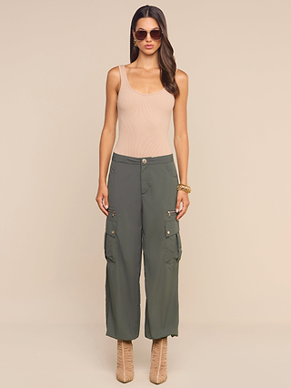 A woman wearing sunglasses, a beige tank top, and the Greer Nylon Jogger in ash olive green with practical cargo pockets stands against a plain beige background. She completes her look with tan lace-up boots, a gold bracelet, and long wavy hair.