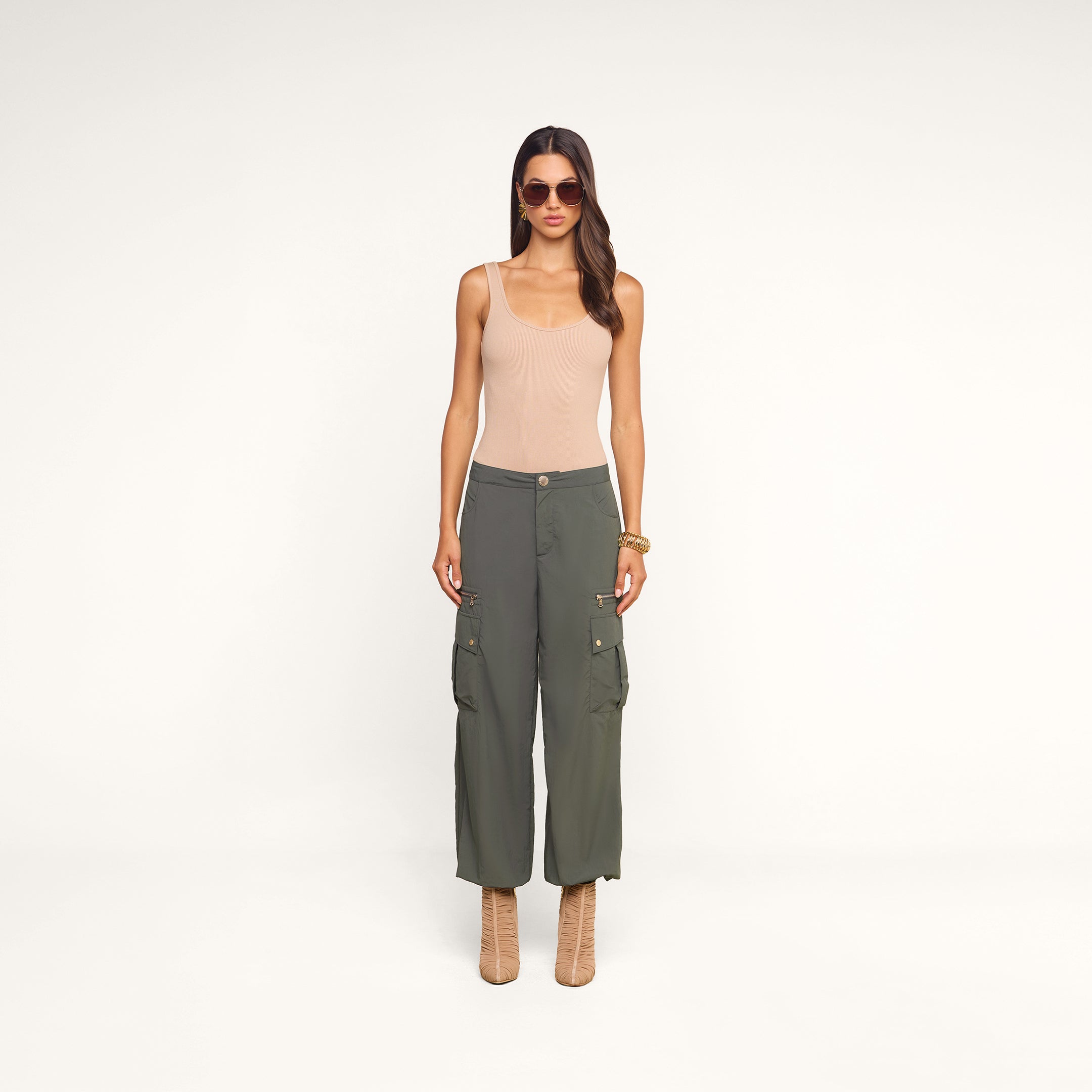 A woman wearing sunglasses, a beige tank top, and the Greer Nylon Jogger in ash olive green with practical cargo pockets stands against a plain beige background. She completes her look with tan lace-up boots, a gold bracelet, and long wavy hair.