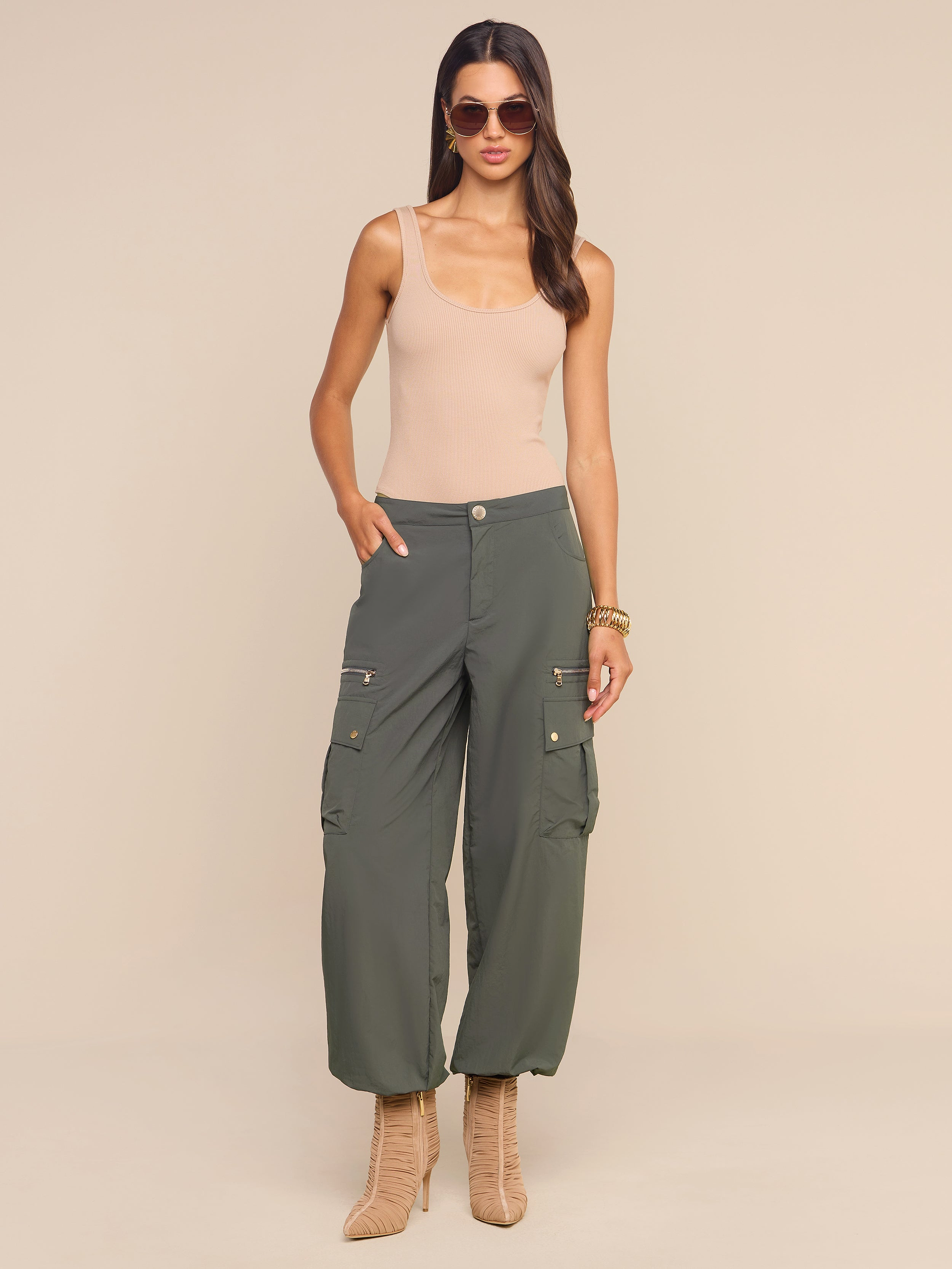 Woman wearing sunglasses, a beige tank top, the Greer Nylon Jogger in ash olive green with cargo pockets, and tan heeled boots stands against a light beige background with one hand in her pocket and the other relaxed at her side.